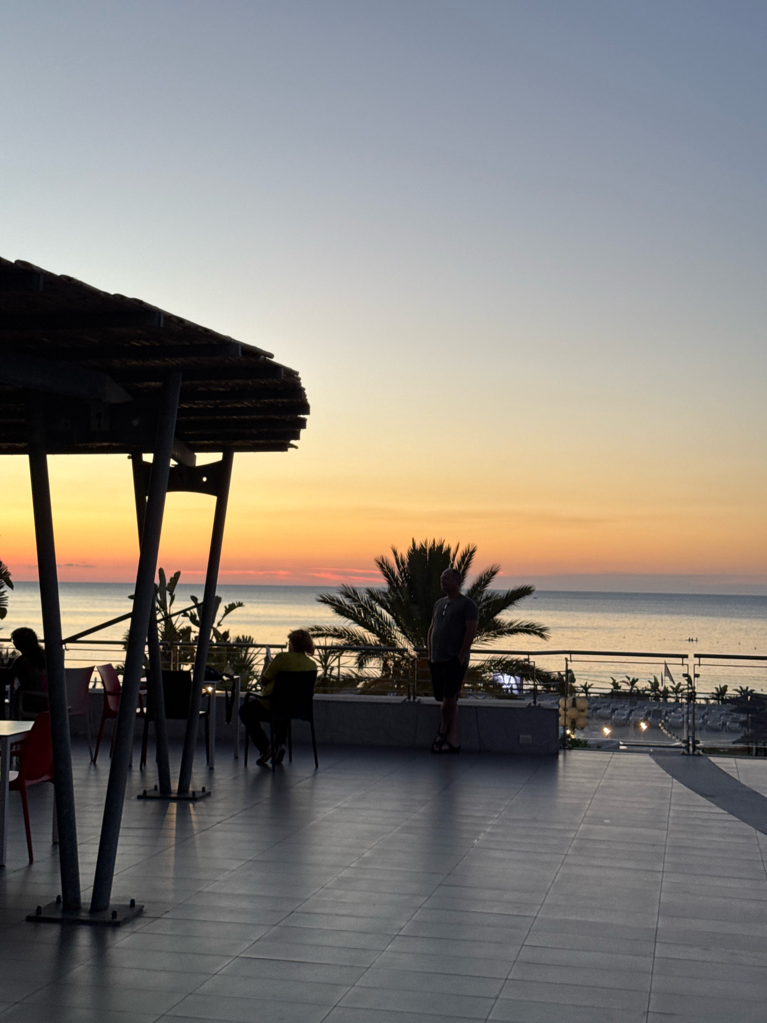 The image depicts an outdoor seating area overlooking the sea at sunset. A thatched roof overhang supported by metal poles covers part of the tiled patio. Several tables and chairs are visible, some occupied by silhouetted figures. In the background, palm trees frame the view of the calm sea, with the sky transitioning through shades of orange and blue. A few people are casually standing or sitting, enjoying the view. In the distance, the coastline and some buildings suggest a populated area. The location is identified as Isola delle Femmine, Italy, indicating a coastal Mediterranean setting.