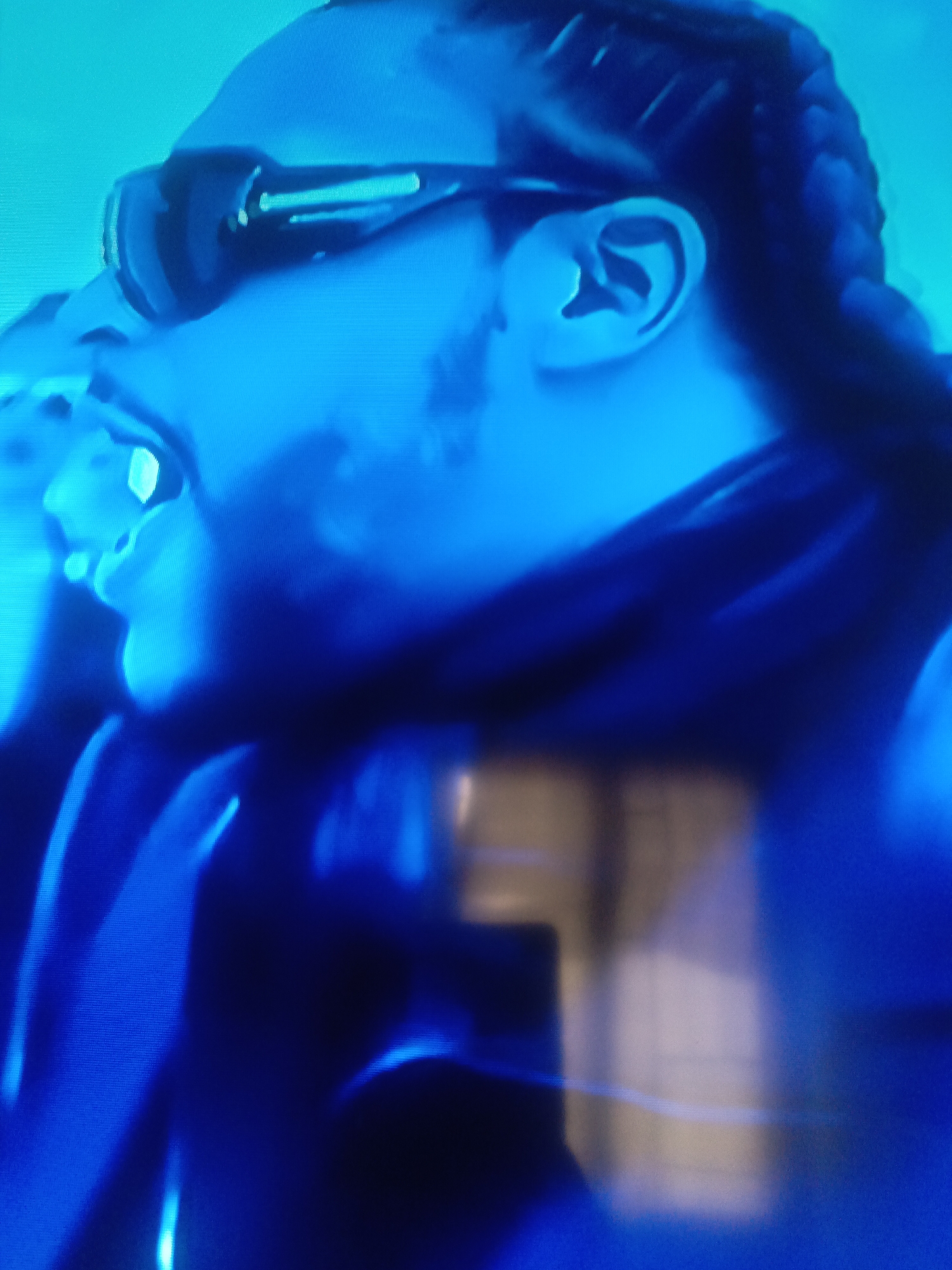 A close-up, low-angle shot in blue lighting shows a person wearing sunglasses and braids. The person is positioned in profile, with their mouth open. Part of their neck and shoulders are visible, clothed in what appears to be dark fabric. The background is indistinct. There is no specific information from the image which indicates it was taken in Juan Griego, Venezuela.