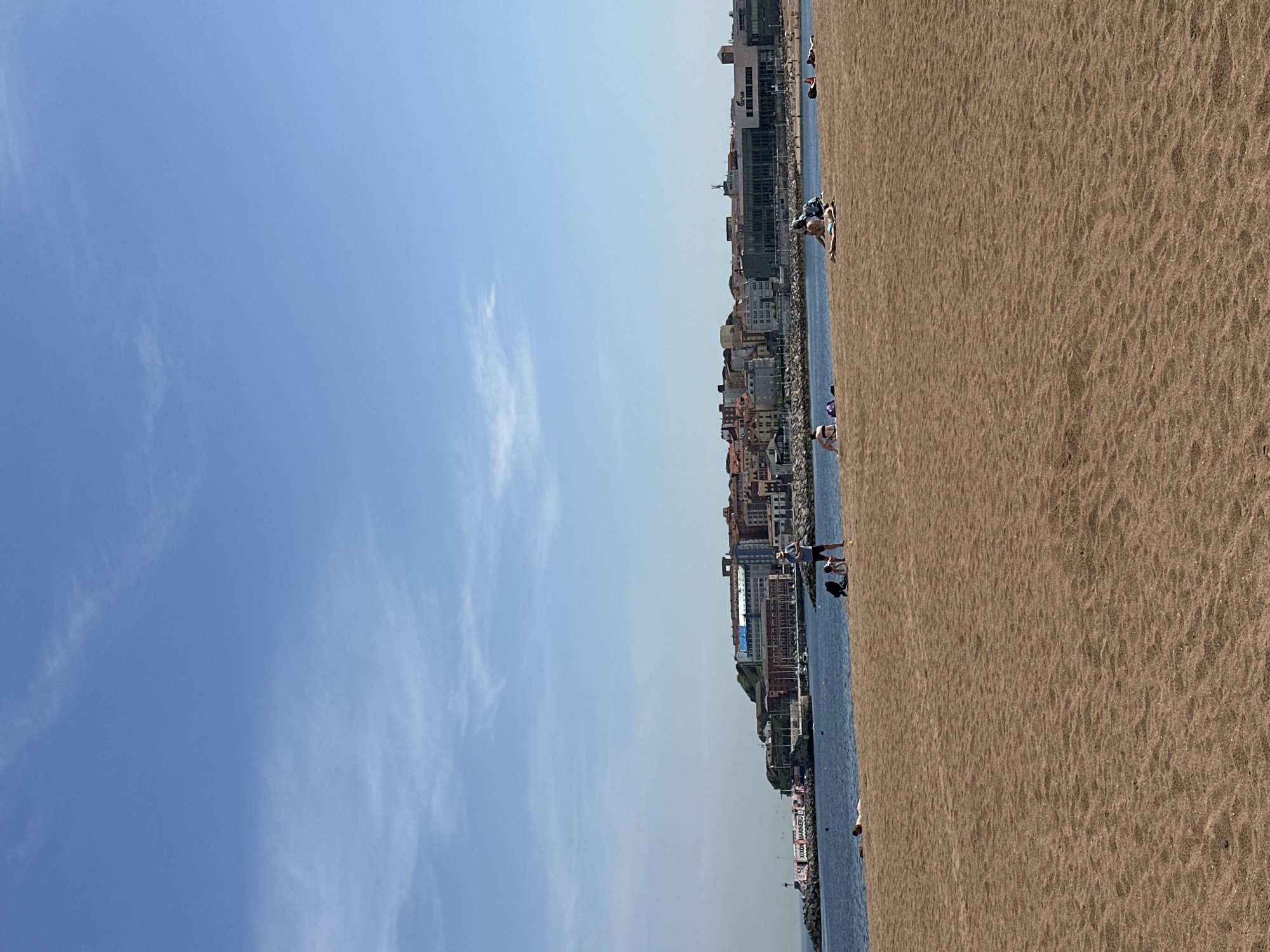 The image shows a beach scene in Gijón, Spain. The beach is sandy, with a shoreline bordering the ocean. A city skyline runs parallel to the beach, featuring buildings of various sizes and architectural styles. People are present on the beach. The sky is blue with scattered clouds.