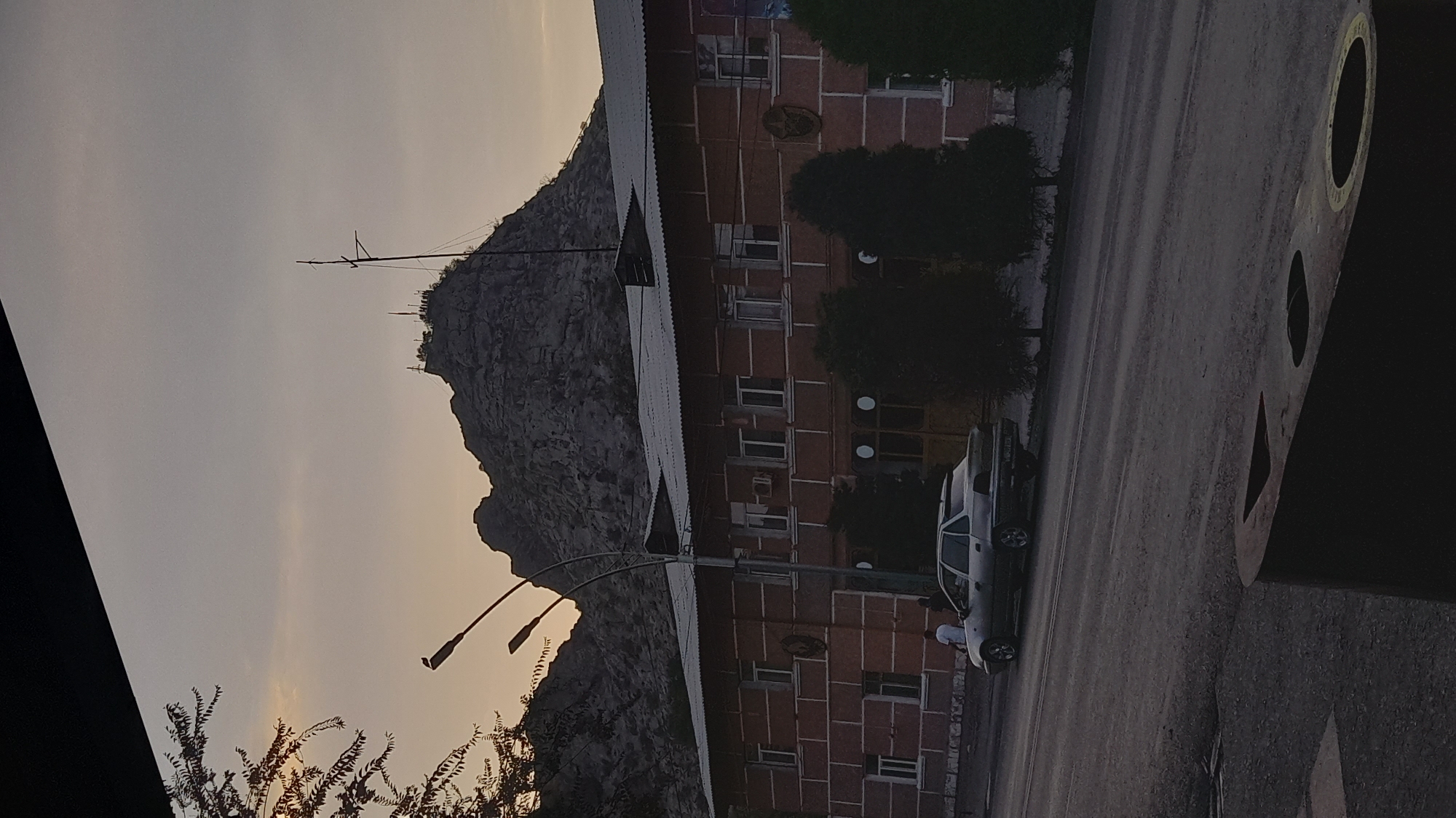 A brick building with white trim and a white roof is situated in front of a rocky mountainside. The building has multiple windows and a few people are visible near a silver car parked on the road in front of the building. The sky is a pale orange and grey, suggesting dusk or dawn. Trees are planted in front of the building. The foreground shows a dark, textured surface with circular cutouts, possibly part of street furniture or infrastructure.