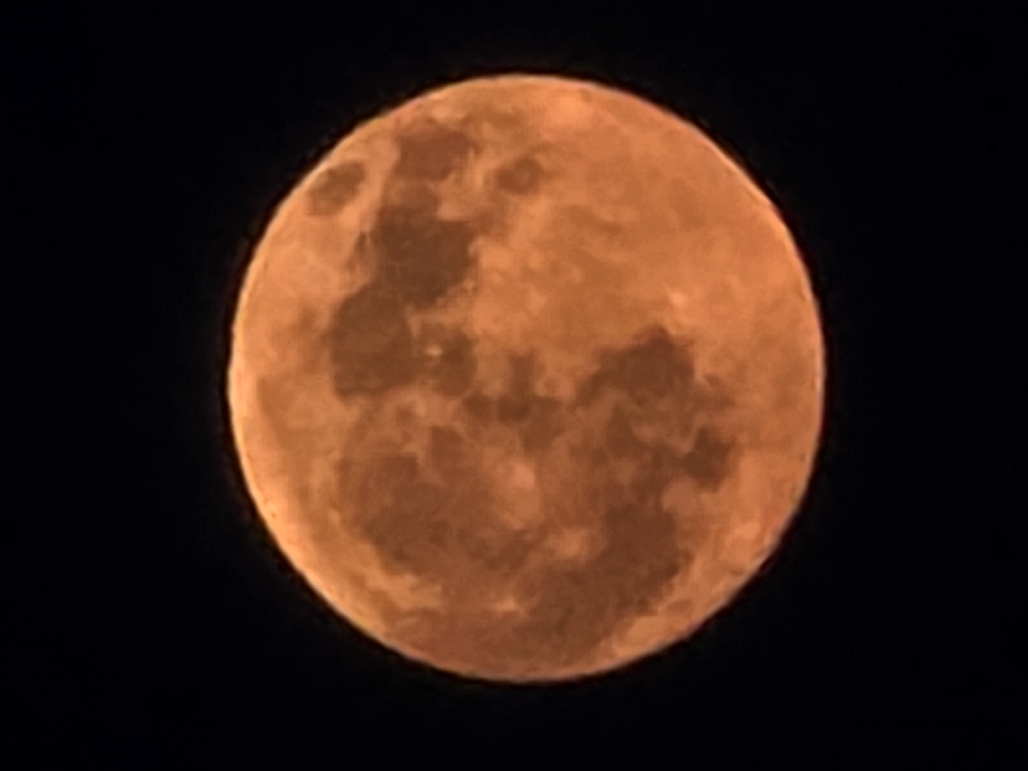 The image is a close-up, vertically oriented shot of a full moon against a black sky. The moon is illuminated with a warm, orange hue, suggesting it is either a harvest moon or the moon is low in the sky and its light is being filtered through atmospheric particles. Surface details of the moon, such as craters and maria, are visible as variations in shade and texture across its disc. The image does not contain any elements that would indicate a specific location in the city of Yambol, Bulgaria, or any human presence.