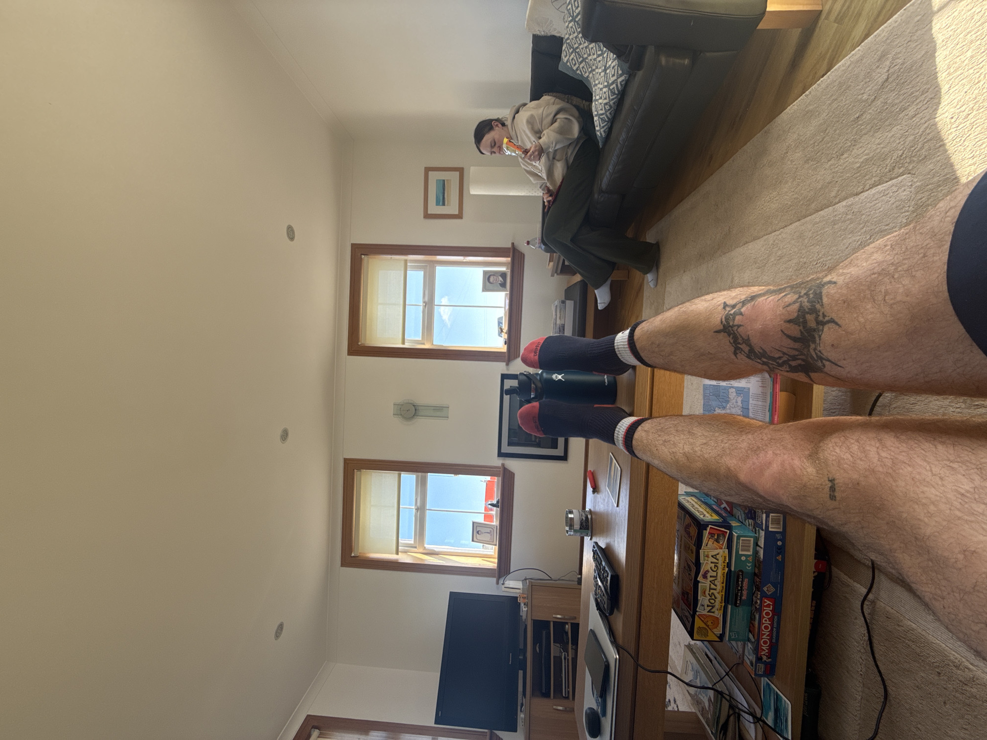 The interior of a living room is depicted in a high-angle, eye-level shot. A person is seated on a dark-colored sofa, eating something from a package. A table, partially visible in the foreground, holds various items, including water bottles, board games, and electronic devices. The foreground is dominated by a person's legs, adorned with tattoos, wearing dark socks with red accents. Natural light streams through windows, illuminating the scene. The location is in a city of Unknown, Unknown.