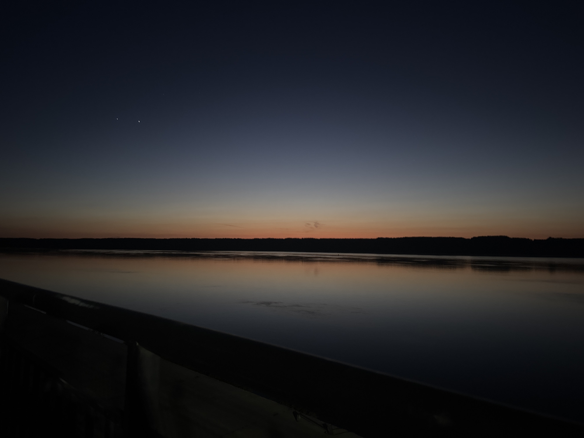 The image depicts a tranquil twilight or predawn scene over a large body of water, likely the Danube River, observed from Lom, Bulgaria. The sky transitions from a band of soft orange and peach hues at the horizon to a deep indigo and dark blue overhead. Two distinct, crescent-shaped celestial bodies, likely the Moon and Venus, are prominently positioned in the upper left quadrant of the sky, with the brighter object slightly to the left and marginally higher than the fainter one. Scattered, very faint stars are also discernible in the darker expanse of the sky.
A dark, silhouetted treeline or forested landmass forms a continuous, low-lying horizon line against the illuminated sky. The calm surface of the expansive body of water reflects the varying tones of the sky, showing darker blues and faint oranges, creating a mirrored effect. Subtle, localized ripples or disturbances are visible on the water's surface in the mid-ground. In the immediate foreground, a dark, out-of-focus structure, resembling a railing or a part of an embankment, extends diagonally from the lower left towards the mid-right, partially framing the water. No individuals, animals, or specific activities are present within the frame.