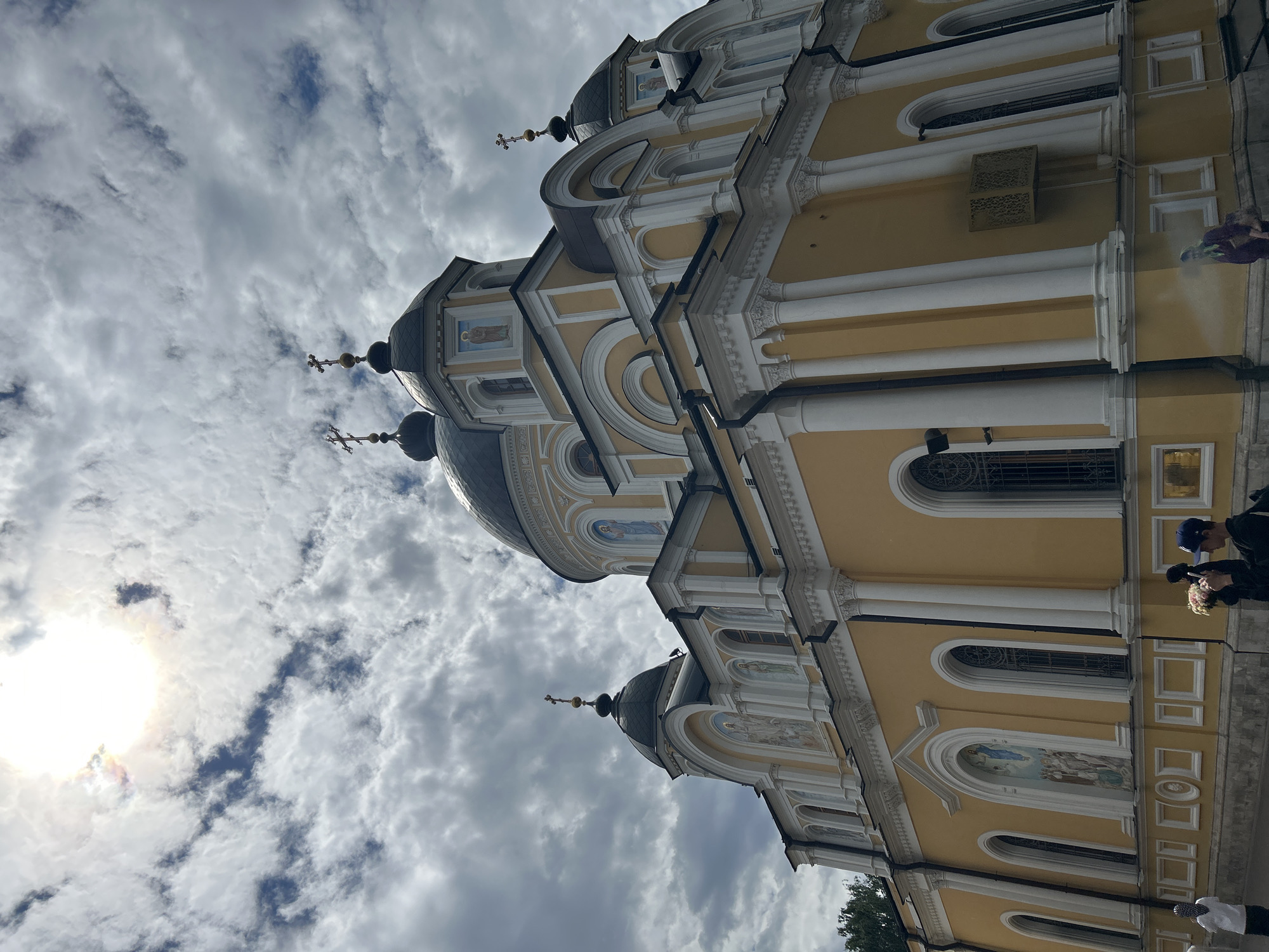 The image displays the exterior of a yellow church with dark domes, possibly an Orthodox church, under a cloudy sky. The sun is visible, peeking through the clouds, suggesting it is daytime, likely mid-morning or afternoon. The architecture features classical columns, arched windows, and ornate details. A cross with a sphere sits atop one of the domes.
Several people are visible in the lower right portion of the image. One person, wearing a blue baseball cap, is holding a bouquet of pink flowers. Another person, partially visible, appears to be dressed in dark clothing. The presence of people suggests a public space, possibly a tourist attraction or a place of worship. The overall scene evokes a sense of serenity and historical significance. The weather appears to be partly cloudy, with the sun creating a bright halo effect as it shines through the overcast sky.