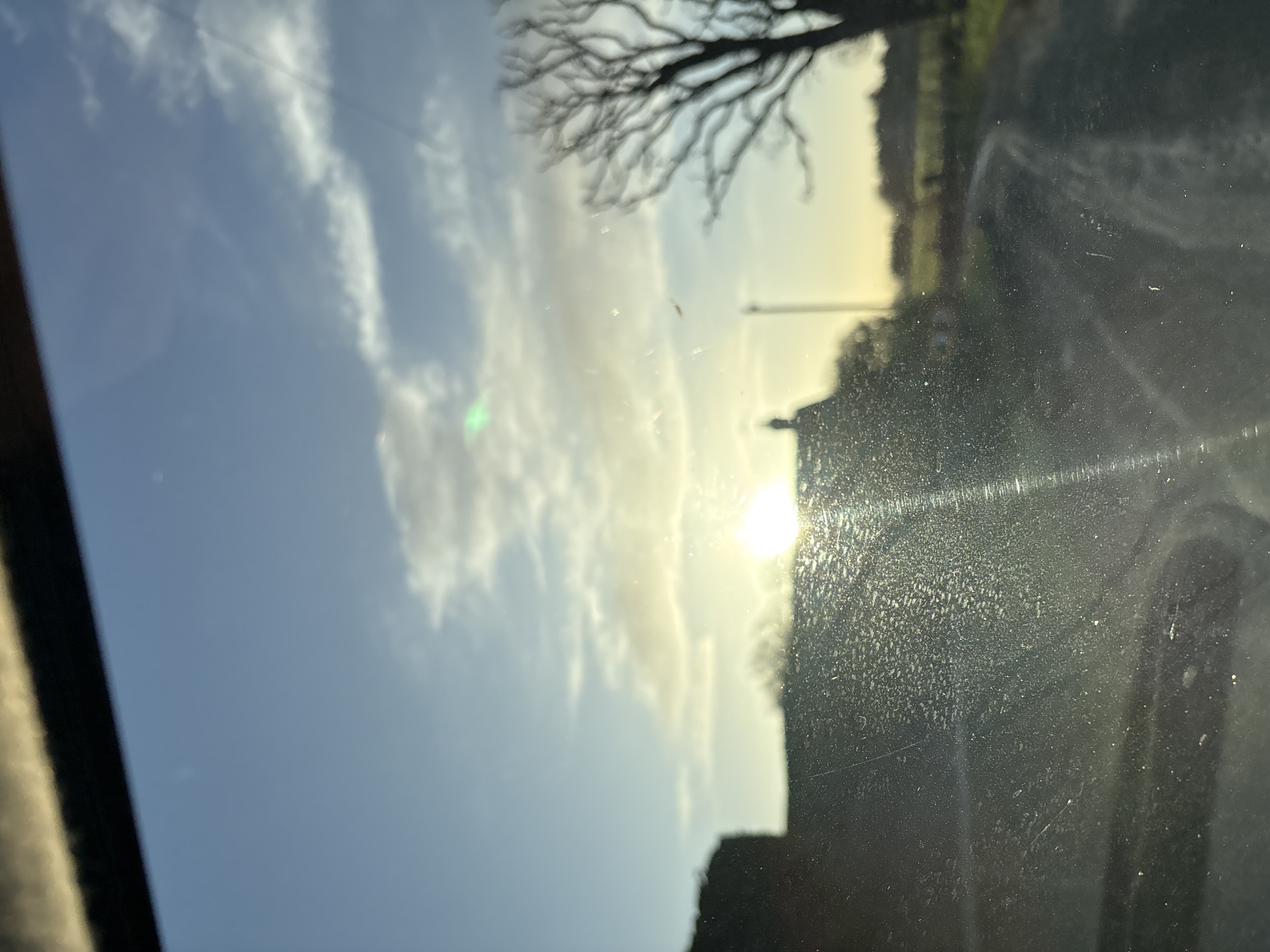 The image presents a rural landscape in Ruyton-XI-Towns, United Kingdom, captured through what appears to be a vehicle's front window. The window surface is notably dirty, covered with a fine layer of dust, small particles, and streaks that interact with light, causing significant glare and lens flare effects, particularly from the prominent sun. A dark, blurred element, consistent with a vehicle's interior trim or dashboard, occupies the top edge of the frame.
The environment outside is dominated by a bright, low-angle sun positioned towards the central-right, suggesting either sunrise or sunset. The sky above is clear to partly cloudy, displaying a light blue hue with scattered white clouds. Silhouetted against the bright horizon are several elements: on the left-center, the dark outline of a building, likely a house, is visible with a discernible roofline and chimney. To the right, a bare, deciduous tree stands with its intricate branch structure. A thin, vertical structure, possibly a utility pole, is faintly discernible between the building and the tree. In the foreground, a dirt or gravel path or road curves from the lower-center towards the right, leading into what appears to be open fields or green land. No individuals, animals, or specific actions are identifiable within the scene.