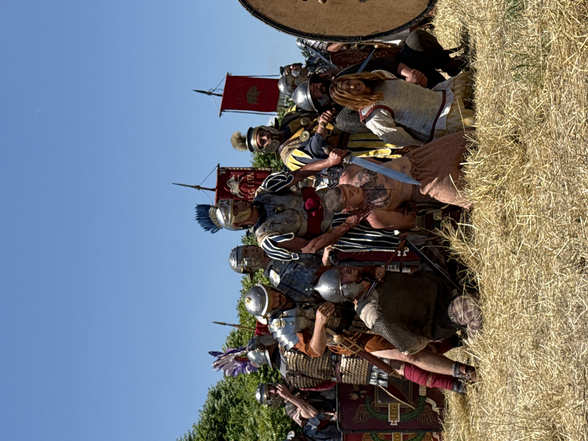 This image captures an outdoor historical reenactment scene in Gigen, Bulgaria, under a clear blue sky with no clouds. The ground is covered with dry, light-brown straw or hay.
In the lower center, two individuals are kneeling. The person on the left is a shirtless male with visible tattoos on his chest and left shoulder, wearing light brown trousers. A Roman soldier's hand, adorned with a red cuff, is placed firmly on his right shoulder/neck area. Adjacent to him, a silver-bladed sword with a blue hilt is partially visible, pointing downwards. The person on the right is also kneeling, wearing a light-colored tunic underneath chainmail armor, and is looking upwards with an open mouth. A Roman soldier's hand rests on their left arm.
Approximately ten individuals dressed as Roman soldiers are visible, primarily standing and observing the kneeling figures. A prominent standing soldier in the central mid-ground wears a silver helmet with a blue transverse crest, ornate silver lorica segmentata armor, a red tunic, and a striped lower garment. His right hand rests on the shirtless kneeling man. To his left (viewer's right), another soldier wears a dark helmet (possibly integrated with chainmail) and chainmail armor, holding a sword. Further to the viewer's left, a soldier with a gold helmet featuring a purple and white plume, and gold scale armor, looks towards the center. On the far right, a large, partially visible round shield stands vertically.
In the upper mid-ground, against the blue sky, two red rectangular Roman military standards (vexilla) are mounted on poles. The standard on the left bears the inscription "LEG.I ITAL," while the standard on the right shows "LEG.I" above an animal emblem, possibly a boar. Green deciduous foliage is visible behind the soldiers, forming a natural backdrop.