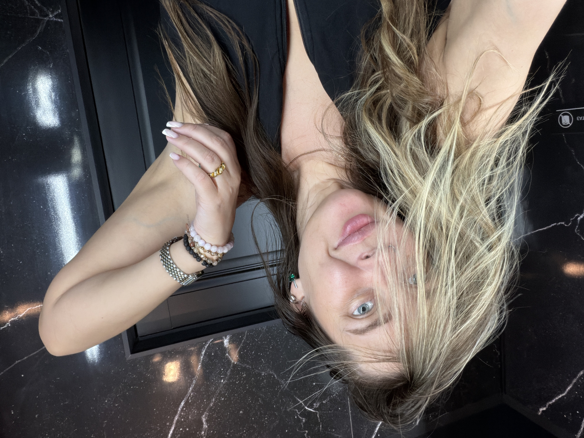 The media file is a close-up, vertically oriented selfie of a young woman with long, wavy blonde and brown hair. She is wearing a black top. Her left arm is bent, and her hand, with manicured nails, is near her face. She is wearing a gold ring with a textured band on her left ring finger and several beaded bracelets and a silver watch on her left wrist. Her right arm is also bent, and her hand is not fully visible.
The woman has blue eyes and light-colored skin. Her hair falls over her face, obscuring parts of it, but her lips are visible, and she appears to be looking towards the camera with a slightly playful expression. She is wearing a small green earring.
The background is dark and reflective, with a marbled pattern. The texture of the background suggests it could be a countertop or a wall. A faint reflection of white light is visible in the upper left of the frame, and a small square icon with rounded corners is visible on the right side, possibly indicating an elevator button or similar control panel. The lighting is artificial and appears to be coming from overhead, creating highlights on her hair and skin. There is no discernible time of day or weather information. The location cues do not provide specific details beyond what is visible in the background. There is no visible text in the image.