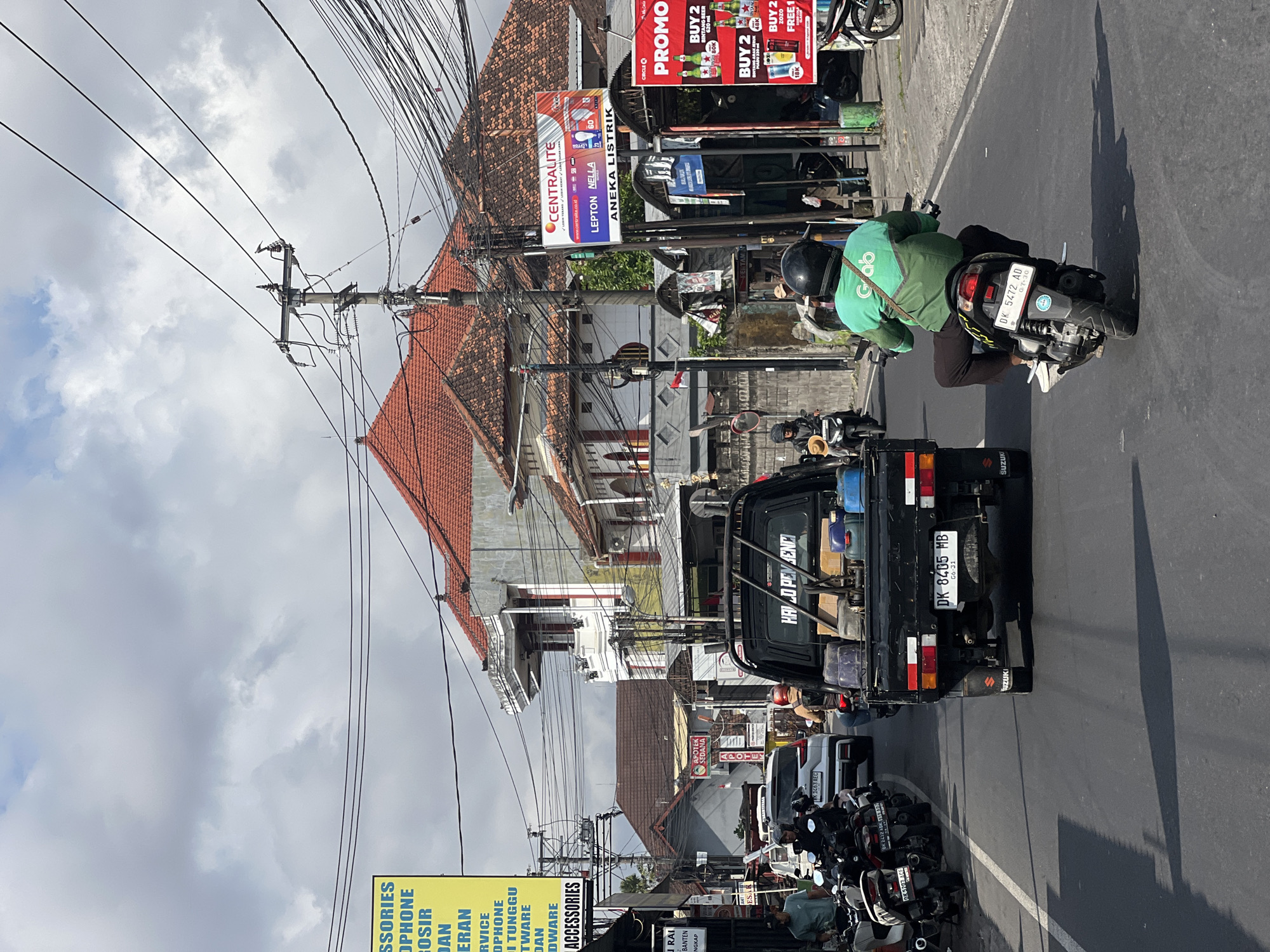 The image depicts a street scene in Denpasar, Indonesia, characterized by a busy road with various vehicles and pedestrians. A black pickup truck with the license plate "DK 8405 MB" is prominently featured in the foreground, carrying various items in its bed. A motorcyclist wearing a green Grab uniform is riding a scooter behind the truck. On the left side of the road, several parked motorcycles are visible. Buildings with red tile roofs line the street, and a large yellow billboard with text in Indonesian is also present. Overhead, a network of electrical wires crisscrosses the sky, suspended from utility poles. Advertisements for Centralite and a "PROMO" are displayed on signs. The sky is partly cloudy, suggesting daytime.
