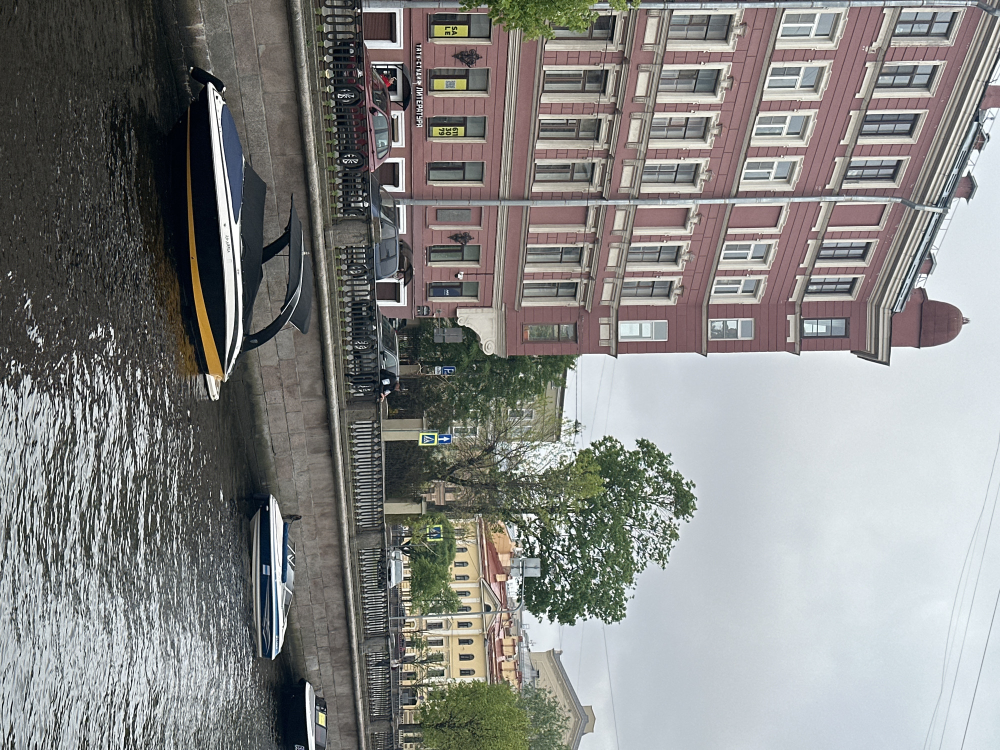 The image depicts a canal in Saint Petersburg, Russia, with several boats docked along a stone embankment. The water in the canal is rippled and dark. The boats are modern, with varying designs and color schemes. A wrought-iron fence runs along the edge of the embankment.
Across the canal, a multi-story building with a distinct red façade and white window frames dominates the background. The building exhibits classical architectural details. Trees with green foliage are visible between the building and the embankment, adding a touch of nature to the urban scene. Cars are parked along the street adjacent to the building. The sky is overcast and grey.