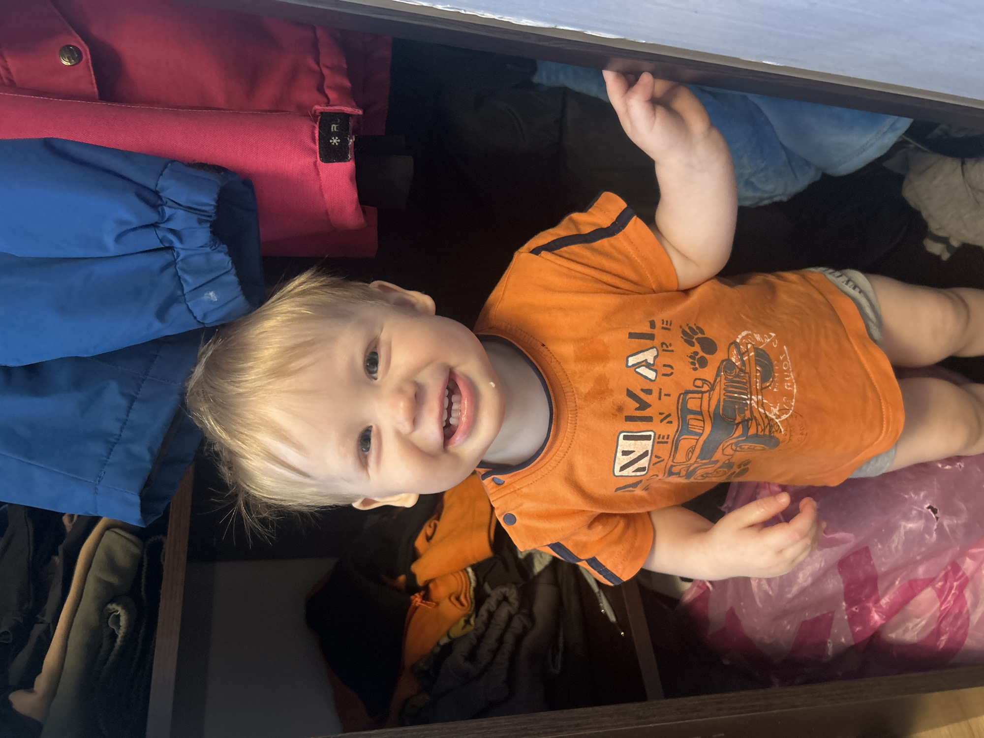 A young child with blond hair and an orange shirt with a vehicle graphic is smiling and looking at the camera. The child appears to be standing inside of an open drawer or shelf. Items of clothing, including a pink jacket and blue jacket, are visible around the child. There is also a translucent pink bag at the bottom of the frame. The location is unknown.