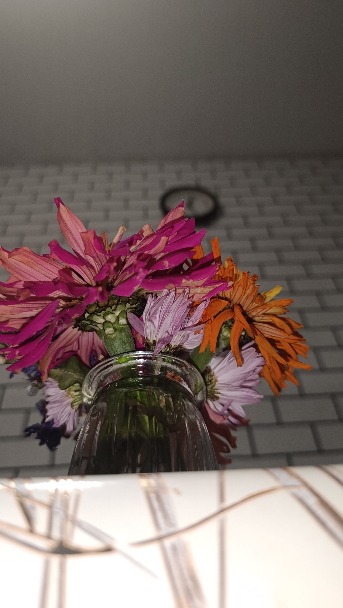 A bouquet of wilting flowers, including pink zinnias and orange chrysanthemums, is displayed in a clear glass vase. The vase sits on a white surface with a patterned design. In the background, a wall with white brick-like tiling is visible, and a circular clock is mounted on it. The scene appears to be indoors, possibly a domestic setting. The lighting suggests it is evening or nighttime. There is no indication of the city of Chaikovsky, Russia, within the image.
