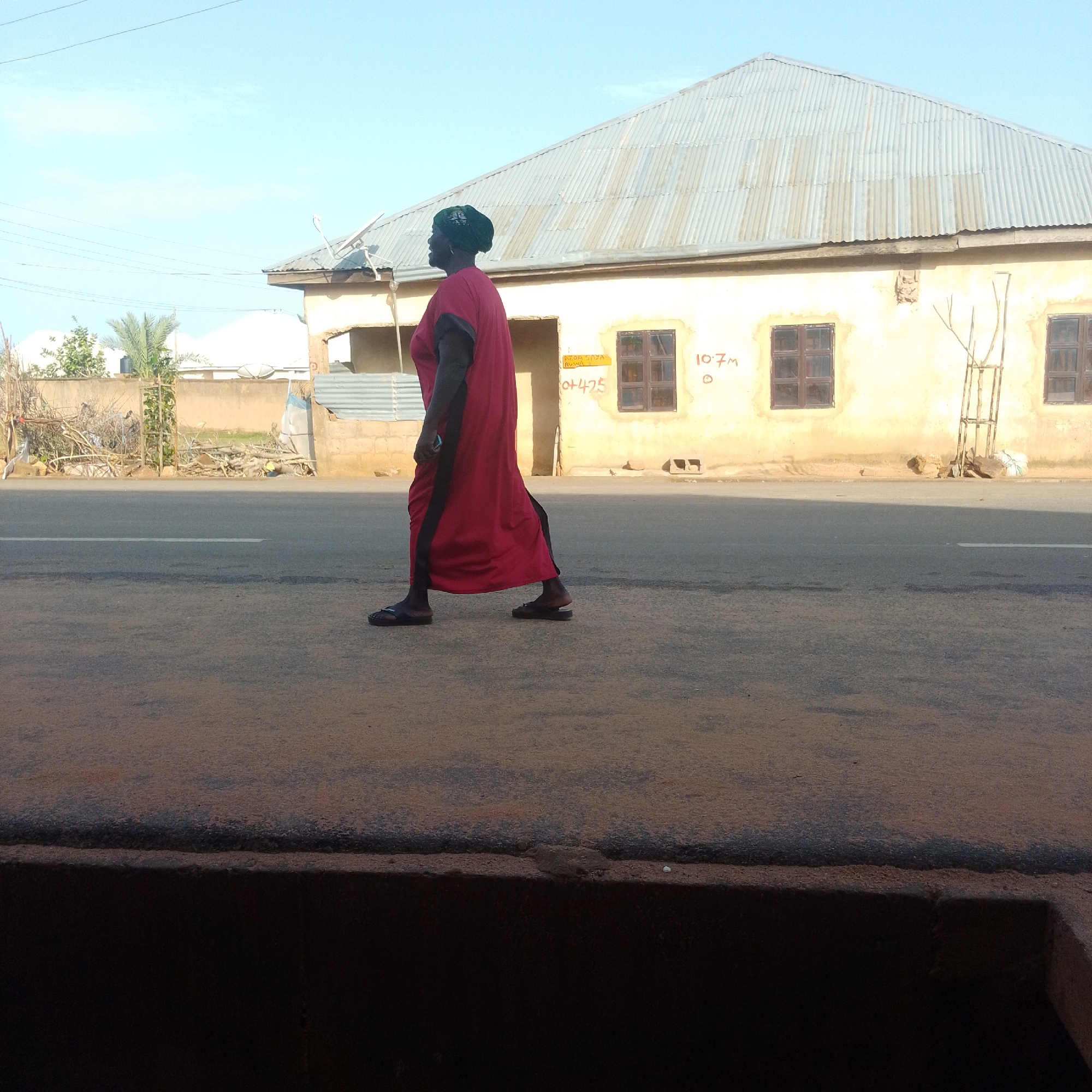 In this media file, a woman walks along a paved road in what appears to be a rural or semi-urban area. She is wearing a long, flowing red dress with a black trim and a dark headscarf. She is walking from left to right across the frame, her face turned to the left, suggesting she is looking in that direction. The woman is wearing dark sandals.
Behind her, a single-story building with a corrugated metal roof stands. The building's walls are a faded yellow, and it has two dark-framed windows visible on this side. There is some handwritten text on the building's wall; in yellow, near one of the windows, it appears to say "10.7M" and below that, "O+475". There's also some smaller yellow text above "O+475". A small, bare tree in a makeshift wooden frame stands to the right of the windows. To the left of the building, there are piles of debris and some vegetation, including a palm tree.
The scene is lit by what appears to be natural daylight, likely late afternoon or early morning given the shadows and the angle of the light. The sky is clear and light blue. The road is asphalt, with a white line marking the lane. The foreground shows a dark, textured surface, possibly a curb or a shallow ditch. The overall impression is one of a typical day in a Nigerian town or village. The woman's expression is not clearly discernible, but her gait suggests purpose.