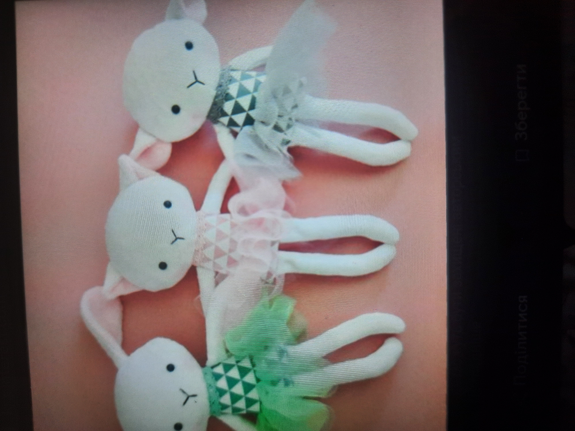 The image shows three white, plush stuffed animal toys, possibly intended to be bunnies or lambs, arranged in a line against a pink background. Each toy has black button eyes and a small "Y" shape embroidered for a mouth. They are all wearing similar tutu-style dresses made of sheer fabric. The leftmost toy wears a green dress with a geometric pattern on the bodice. The middle toy wears a pink dress with a similar geometric pattern. The rightmost toy wears a white dress with a dark grey and white geometric pattern on the bodice. The stuffed toys are positioned from left to right, and their legs are hanging down. The bottom of the image shows a dark surface with text that appears to be in Russian, reading "Поделиться" (Share) on the left and "Сберегти" (Save) on the right. There is no information in the image that indicates a location in the city of Rosenheim, Germany.