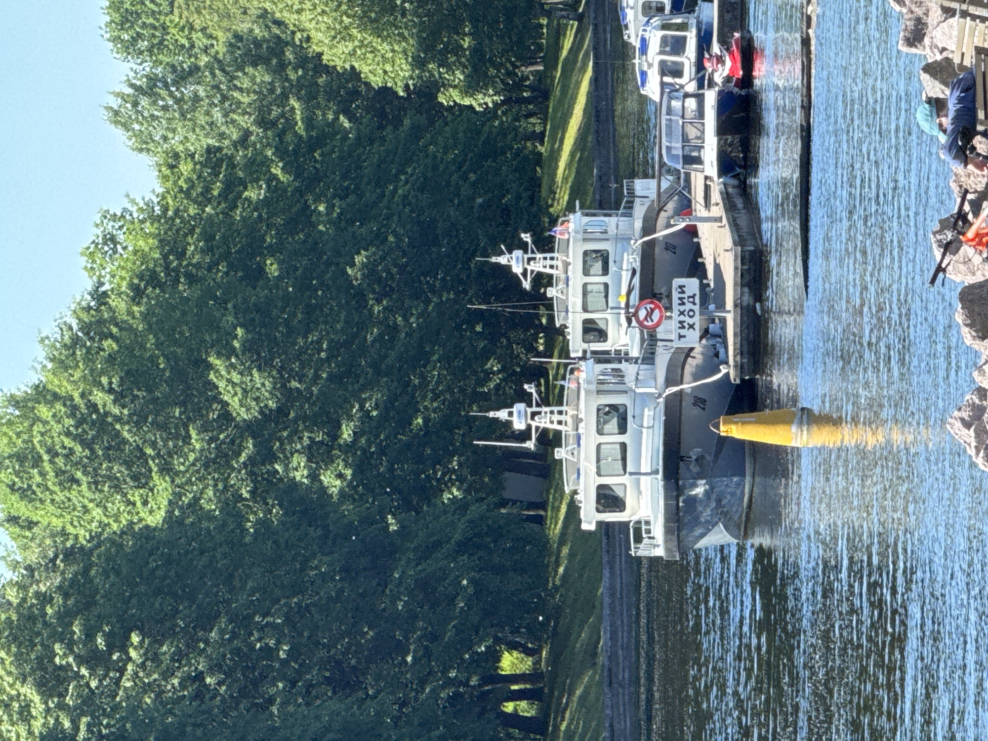 The image depicts a scenic canal in Saint Petersburg, Russia, likely within the city's extensive network of waterways. The canal is flanked by a lush, green embankment covered in dense trees, suggesting a park or natural area bordering the water. The sky above is a clear, bright blue, indicating fair weather.
In the foreground, the calm blue water reflects the sunlight, creating a shimmering effect. Moored along a wooden pier are several vessels. The most prominent are two white boats, appearing to be patrol or service boats, with enclosed cabins and radar equipment on top. One of these boats has the number "218" visible on its hull. A yellow, conical buoy floats in the water near the pier.
Attached to the pier is a sign with a red circle and a diagonal line across a symbol, indicating a prohibition, and text below that reads "ТИХИЙ ХОД" (TIKHIY KHOD), meaning "Slow Speed" or "No Wake."
To the right of the main boats, a smaller blue boat is docked, and next to it, a red personal watercraft (jet ski) is visible. Further to the right, another white boat with blue trim is partially seen.
In the lower right portion of the image, a person is seen relaxing on a wooden bench, leaning back and wearing a blue cap. A bicycle with an orange frame is parked next to the bench. The area around the bench is rocky. The overall scene conveys a peaceful and tranquil atmosphere in an urban waterway environment.