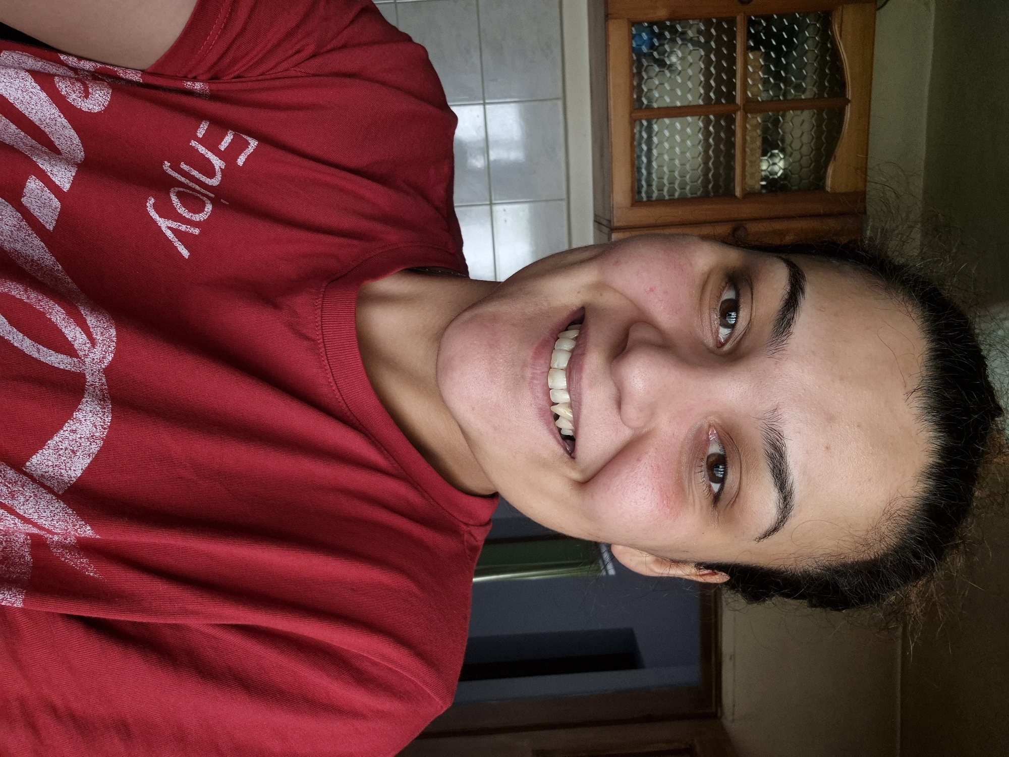 The image depicts a young adult female in a frontal, chest-up shot within an interior room located in Iskrets, Bulgaria. The subject has dark hair pulled back, dark eyebrows, and dark eyes. She is smiling broadly, exposing her upper teeth. Her skin exhibits visible pores and slight redness on the cheeks. She is wearing a short-sleeved, round-neck red t-shirt with white text on the front; the word "Enjoy" is clearly visible, and part of the "Coca-Cola" logo is discernable below it.
The background on the left side features a wooden wall cabinet with glass doors composed of multiple panes, each fitted with textured hexagonal glass. Inside the cabinet, indistinct objects are visible, including a white spherical item in the upper right pane and a blue object in the lower left pane. Below the cabinet, a wall tiled with light-colored square tiles and visible grout lines extends across the frame. The background on the right is darker and less distinct, suggesting a darker wall or doorway. Illumination appears to be from the front, casting soft shadows.
