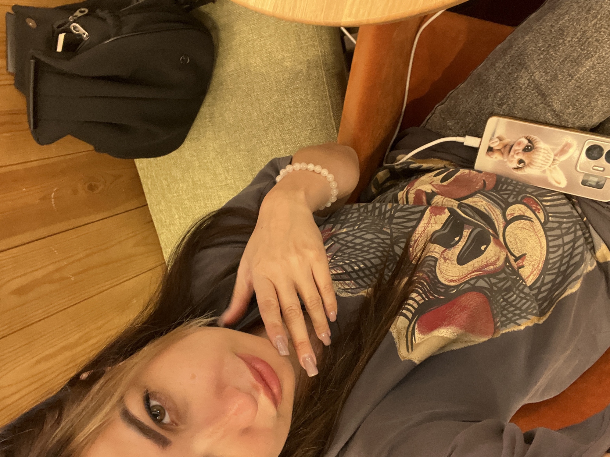 A woman is taking a selfie in an indoor setting. She is wearing a dark t-shirt with a graphic print and gray pants. Her right hand is resting on her chin, and she is wearing a light-colored bracelet on her left wrist. A smartphone, plugged in and displaying a picture of a rabbit, rests on her lap. In the background, a black backpack is visible on a green upholstered seat, and wooden paneling lines the wall. The scene appears to be in an urban environment, though the specific city and location are unknown.