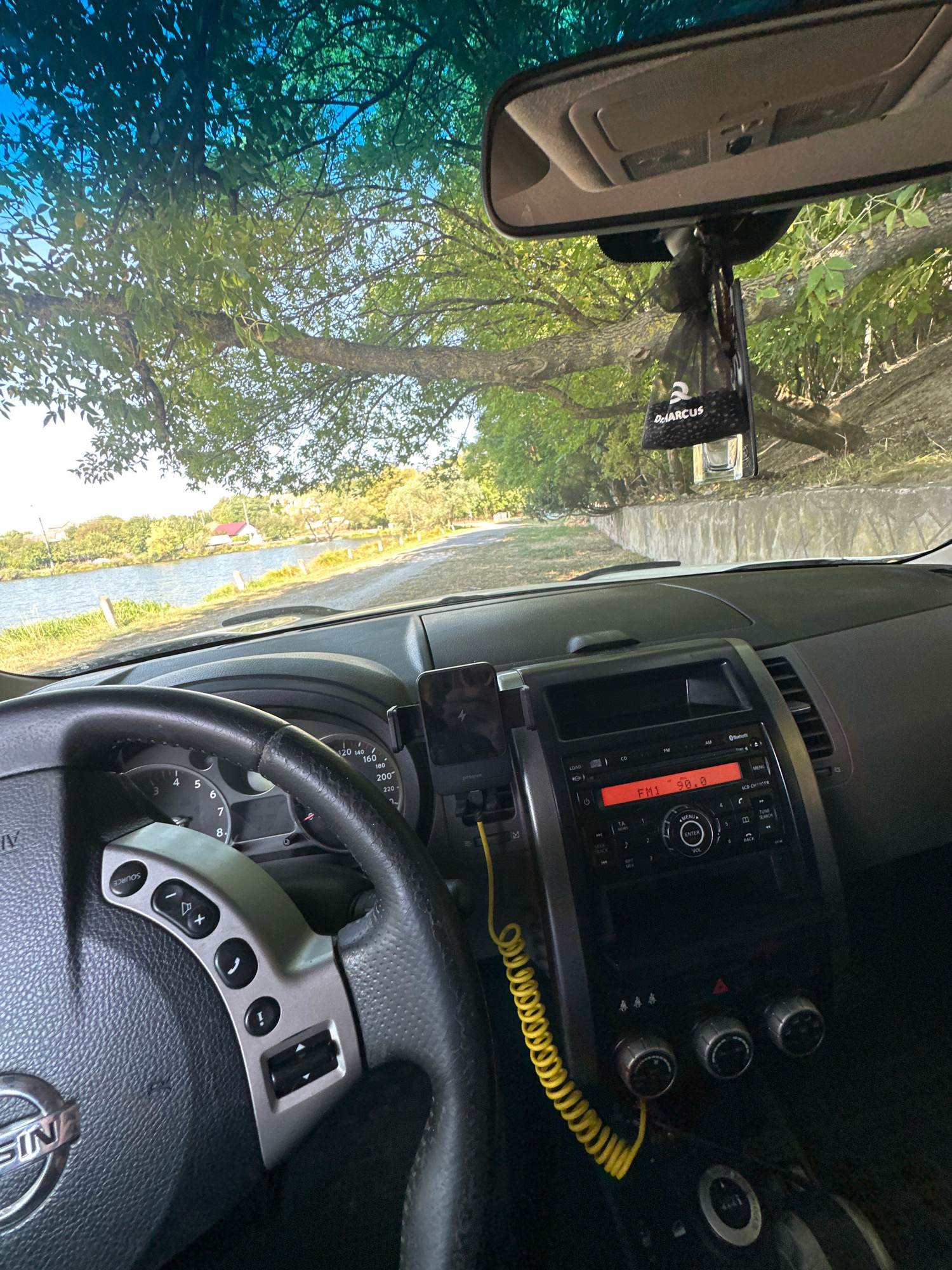 The image captures the interior view of a car parked outdoors, likely near a body of water. Through the windshield, a tree with green leaves overhangs the road. In the distance, a tranquil scene unfolds with a calm body of water, bordered by trees and vegetation. Several houses with red roofs are visible on the far bank. The car's dashboard is prominent, featuring a steering wheel with various controls, a car radio displaying "FM1 96.0", and a smartphone mounted on the dash. A black car air freshener hangs from the rearview mirror. The setting appears to be a serene, rural environment, possibly a lakeside or riverside location. While the image is stated to be in Makiv, Ukraine, this specific location is not visually confirmed within the image itself.