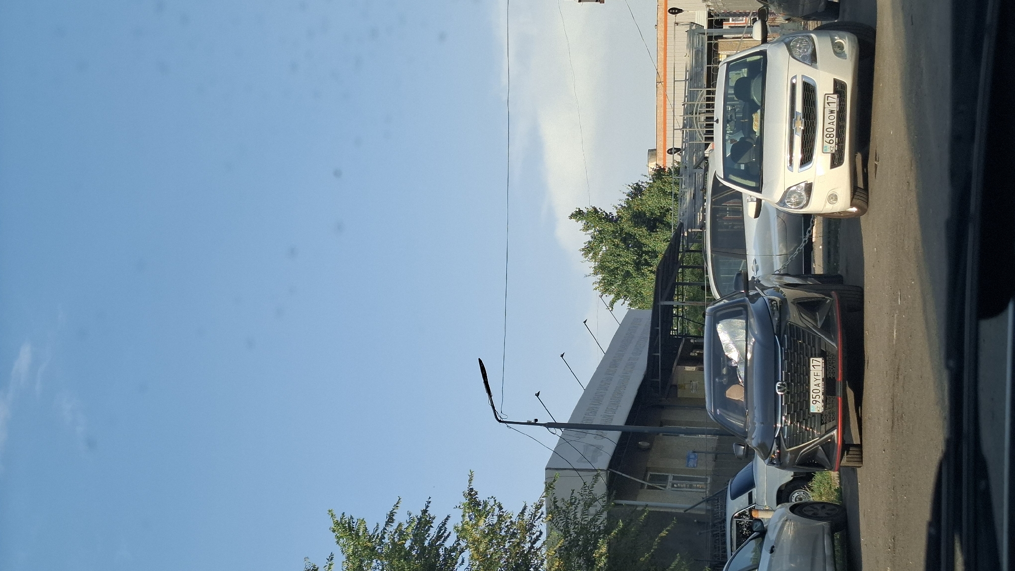 The image depicts a street scene in Shymkent, Kazakhstan, under a clear blue sky with scattered clouds. The foreground shows the hoods of two cars, with a dark gray car on the left and a white Chevrolet on the right. The dark gray car has a license plate reading "KZ 950 AYF 17" and red accents on its front bumper. The white Chevrolet has a license plate reading "KZ 680 AOW 17".
Behind the cars, a building with a white facade and dark windows is visible. A street lamp with wires extends from the left side of the frame towards the building. Trees with green foliage are present on the left and in the background, and another building with an orange-topped structure is seen further in the background on the right. The overall impression is a typical urban environment, likely a parking lot or street in Shymkent.