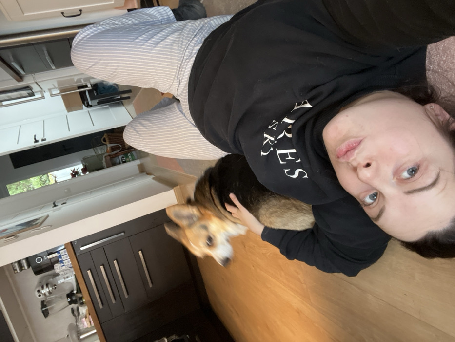 The image displays a female individual and a dog on a light-toned wooden floor within a domestic residence, identified as being in Obermoschel, Germany. The female subject is lying supine, facing the camera with her lips pursed. She wears a black hooded sweatshirt featuring white inverted text on the chest and light-colored, vertically striped pants. Her right hand rests on the flank of a tri-color Pembroke Welsh Corgi, positioned closely beside her. In the background, a kitchen area is visible, comprising dark-colored base cabinets with metallic bar handles, a light countertop, and various appliances including a stand mixer, a blender, and a coffee maker. A white interior door and a partial view of outdoor foliage are also present, along with a woven basket near a potted plant.