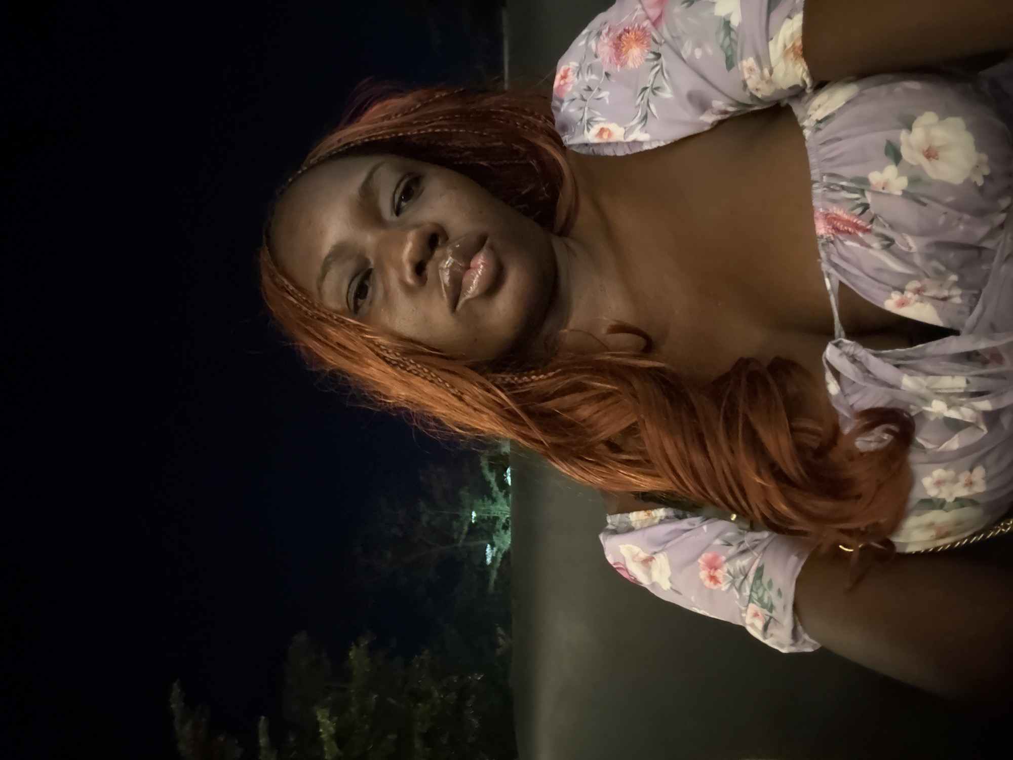 The image shows a woman with auburn braided hair. She is wearing a light purple floral dress. The background is dark, with some green foliage visible, and the image was taken at night. It's not possible to determine if the location is in the city of Abuja, Nigeria, or any other specific location.