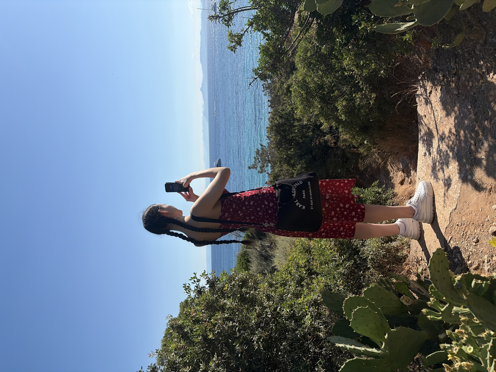 A young woman with braided hair stands on a dirt path overlooking the ocean, holding up a phone to take a picture. She is wearing a red floral dress and white sneakers, and has a black messenger bag over her shoulder. The ocean is a vibrant blue, with a few boats visible in the distance. The path is lined with green bushes and cactus plants. The sky is clear and blue. The image appears to have been taken in a coastal area, possibly a park or nature reserve. The specific location in Vouliagmeni Municipal Unit, Greece, is not discernible from the image itself, but the scene is consistent with a Mediterranean coastal landscape.