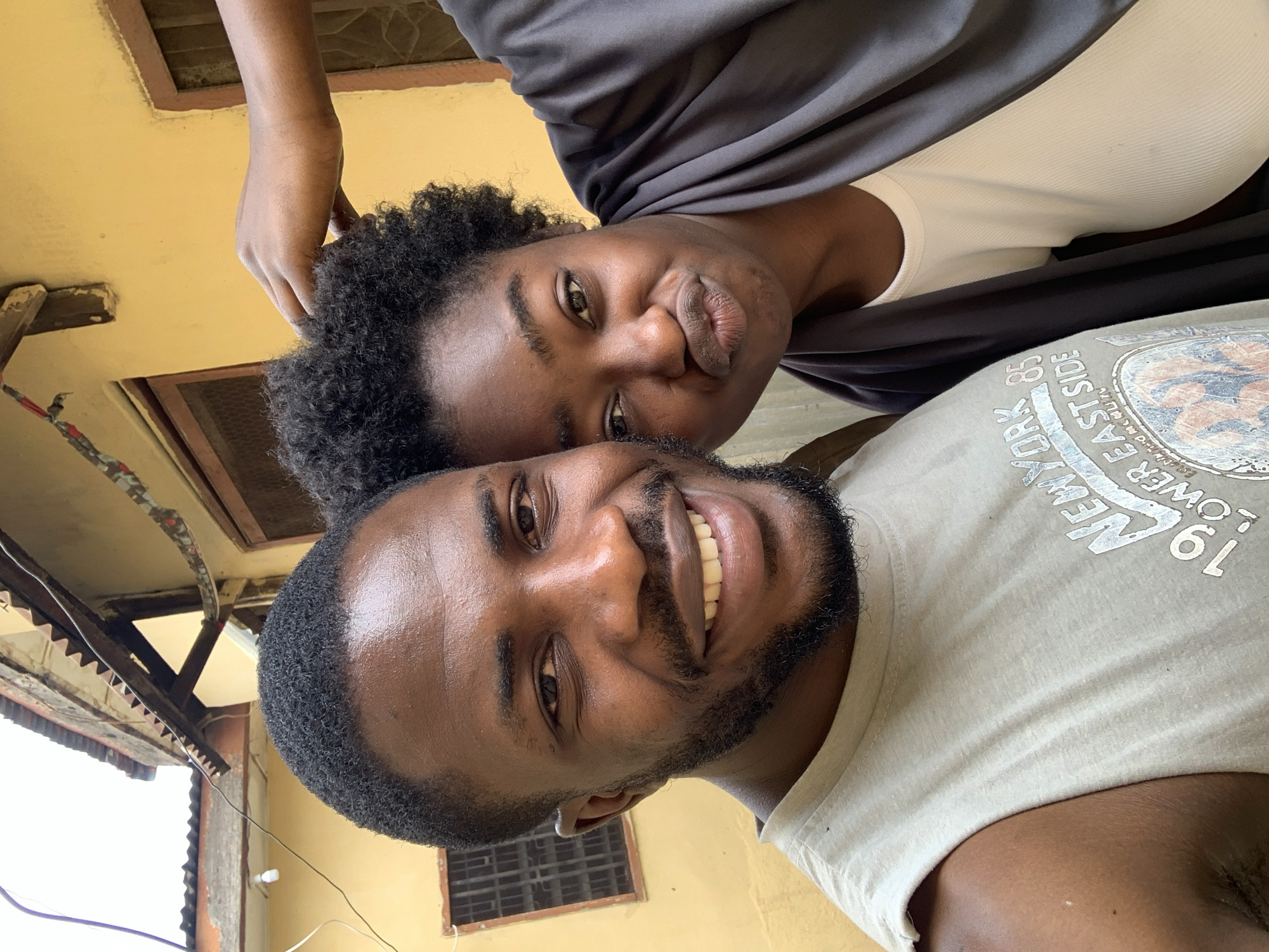 A selfie taken from below depicts two people. The person in the foreground has dark skin, a beard, and is smiling. They are wearing a light-colored t-shirt with a graphic and the words "NEW YORK EAST SIDE" are visible. Behind them is a person with dark skin and short, curly hair, pouting at the camera. They are wearing a white top and draped in a dark fabric. A hand touches the hair of the person in the background. The background shows a yellow building with windows, wiring, and part of a corrugated roof. The location is in Rafin Par, Nigeria.