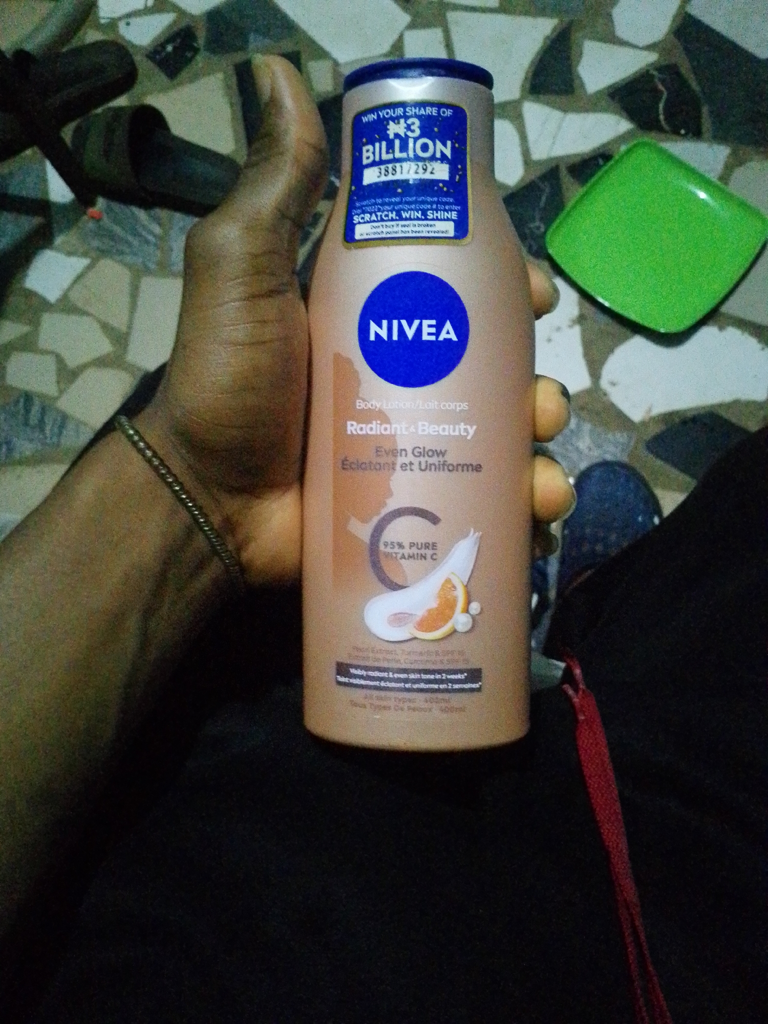 A person holds a bottle of Nivea Radiant + Beauty Even Glow body lotion. The lotion claims to contain 95% pure vitamin C and promises visibly radiant and even skin tone in 2 weeks. A sticker indicates a promotion where one can win a share of ₦3 billion with a unique code revealed by scratching a panel. The background shows a green plastic container and a floor with mosaic tiles. A sandal and a shoe are also visible. The image was taken in Ipetumodu, Nigeria.