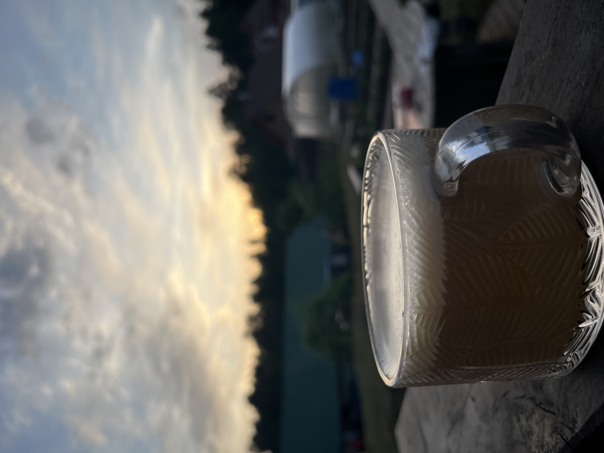 The image depicts a glass mug filled with a light-colored liquid, likely coffee or tea. The mug rests on a dark, wooden surface, possibly a deck railing. In the background, there is a blurred view of a partly cloudy sky tinged with the warm colors of sunset or sunrise. Behind the mug, there is a blurry landscape of trees and a white structure.
The location of the image is not evident from the scene details. The image does not provide any specific details which identify Chaikovsky, Russia.