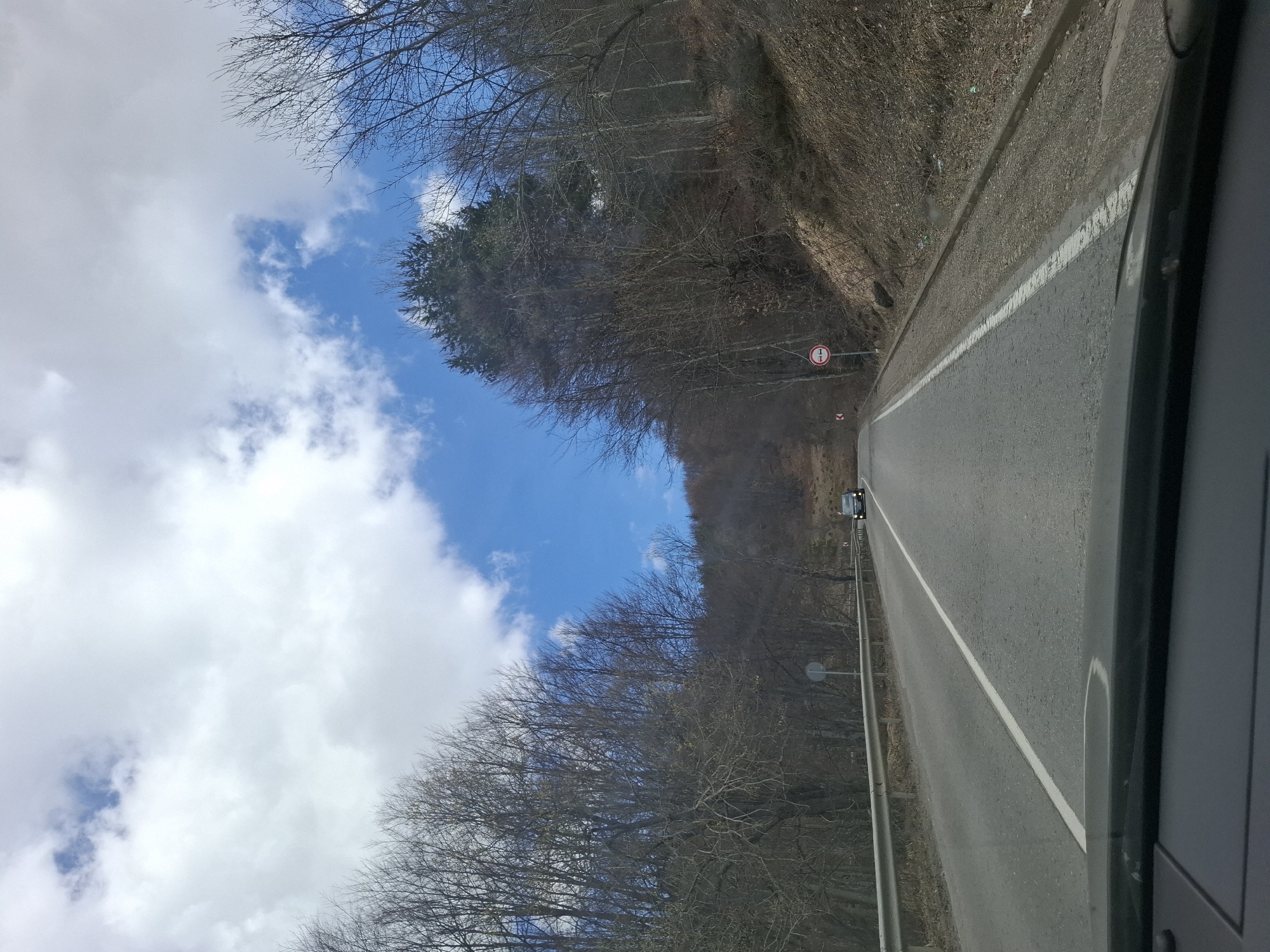 The image shows a view from inside a car, looking down a straight two-lane road. The road surface is gray asphalt with a white line marking the center. A vehicle is visible in the distance on the road.
The road is flanked by a steep, vegetated hillside on the right. On the left, bare trees line the road, with patches of blue sky and white clouds visible above.
A road sign is located on the right side of the road.
The location of the image is not possible to deduce from the image alone.