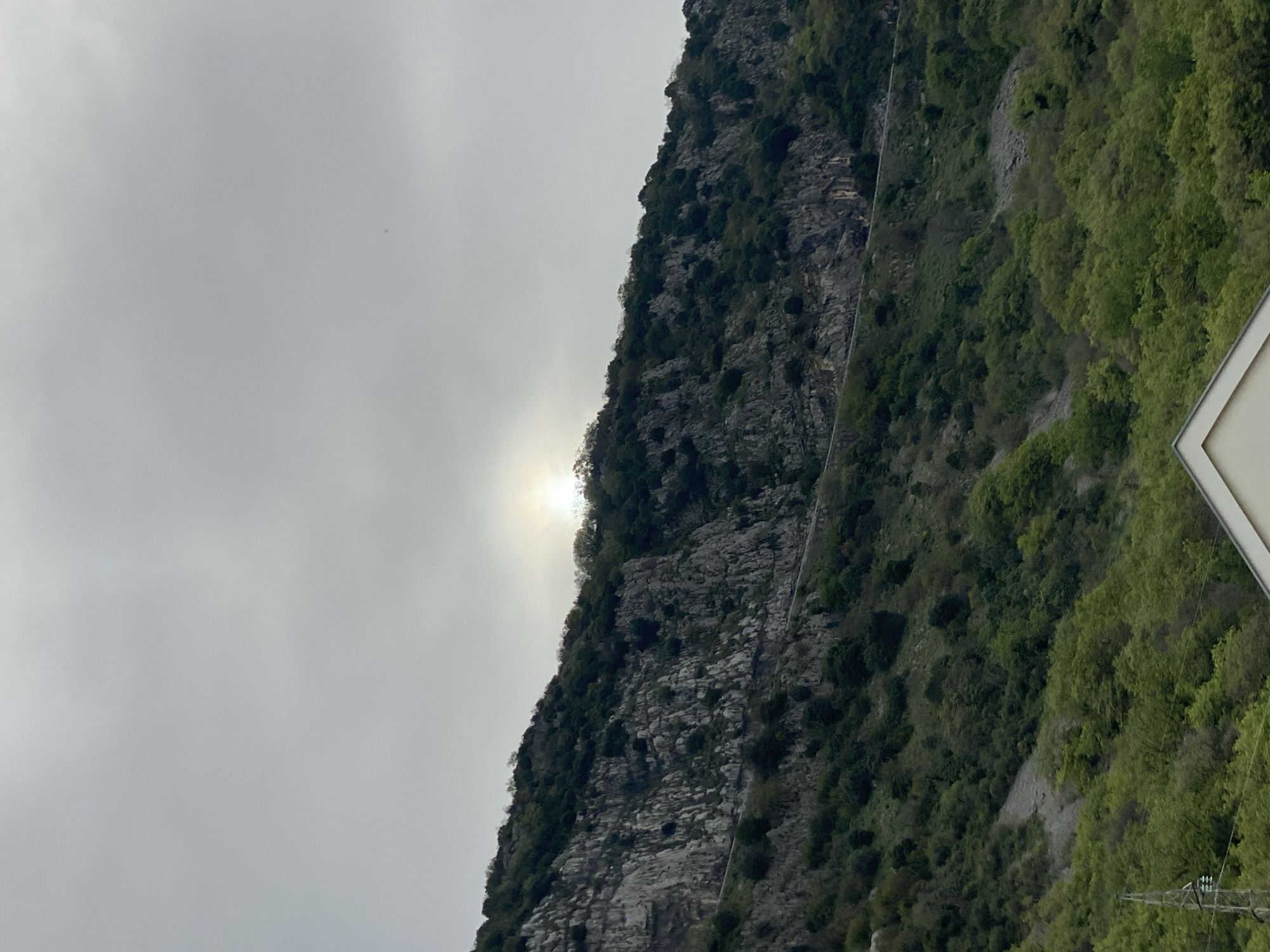 The image shows a view of a rocky mountain covered with trees under an overcast sky. The sun is partially obscured by the mountain's peak, creating a bright glare. A road or path is visible winding along the mountain's side. A section of what appears to be a sign is visible in the lower right corner of the image. Based on the terrain and vegetation, this could potentially be a scene in or around Budva, Montenegro, as the area is known for its mountainous coastal landscapes.