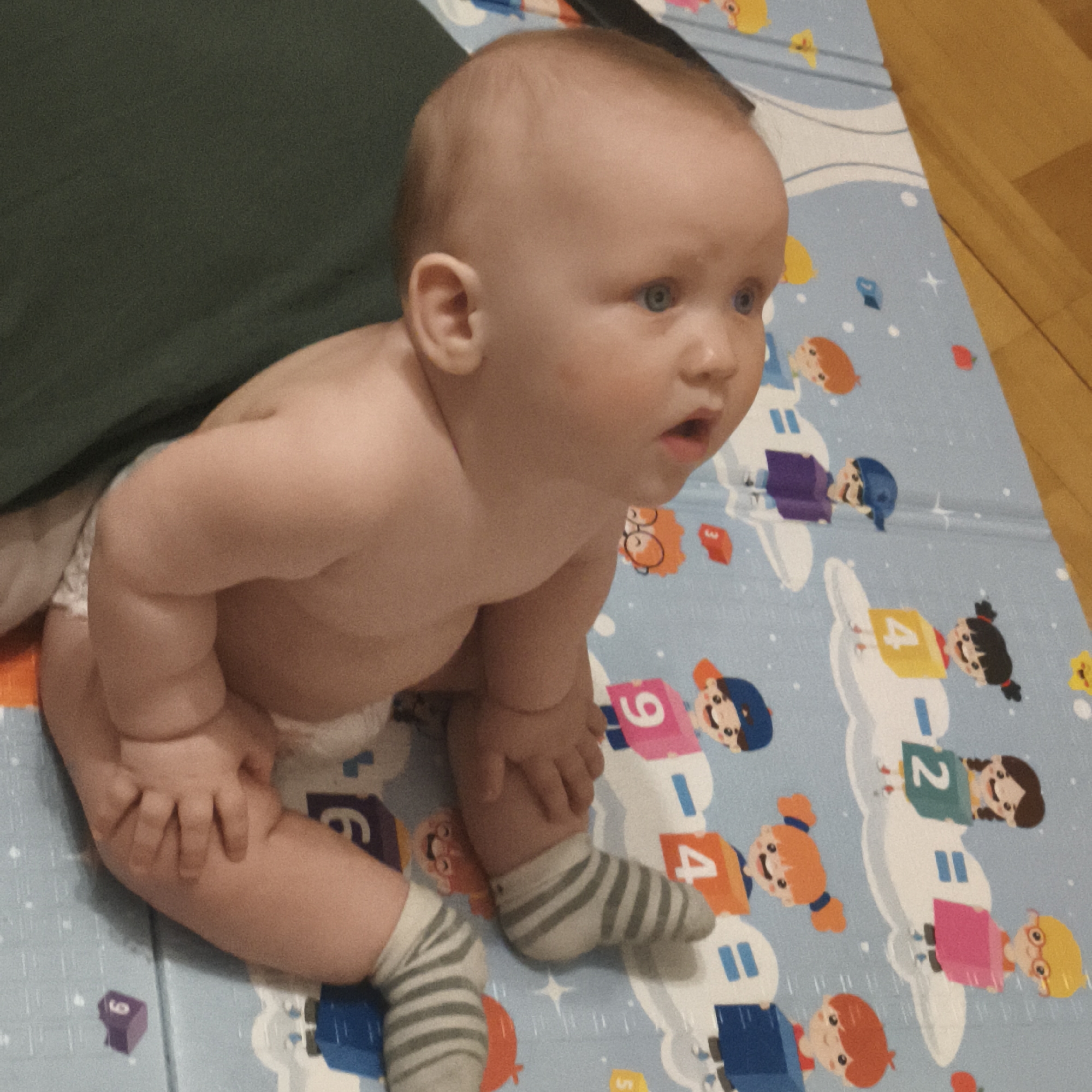 A baby sits on a blue play mat with cartoon figures and numbers. The baby wears a diaper and grey and white striped socks. The baby is partially covered by a dark green pillow or cushion to the left. The floor is wooden.