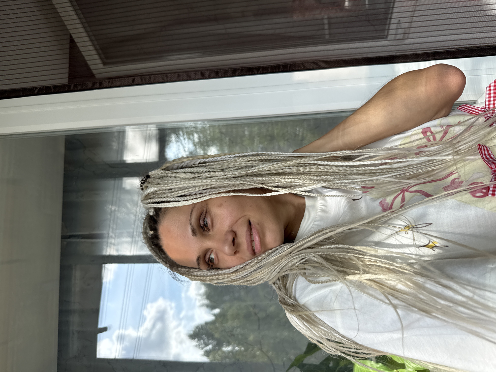 A woman with blonde braided hair, wearing a white t-shirt, stands near a window. The background shows trees and a sky reflected in the glass. The scene appears to be outdoors, possibly on a balcony or patio.