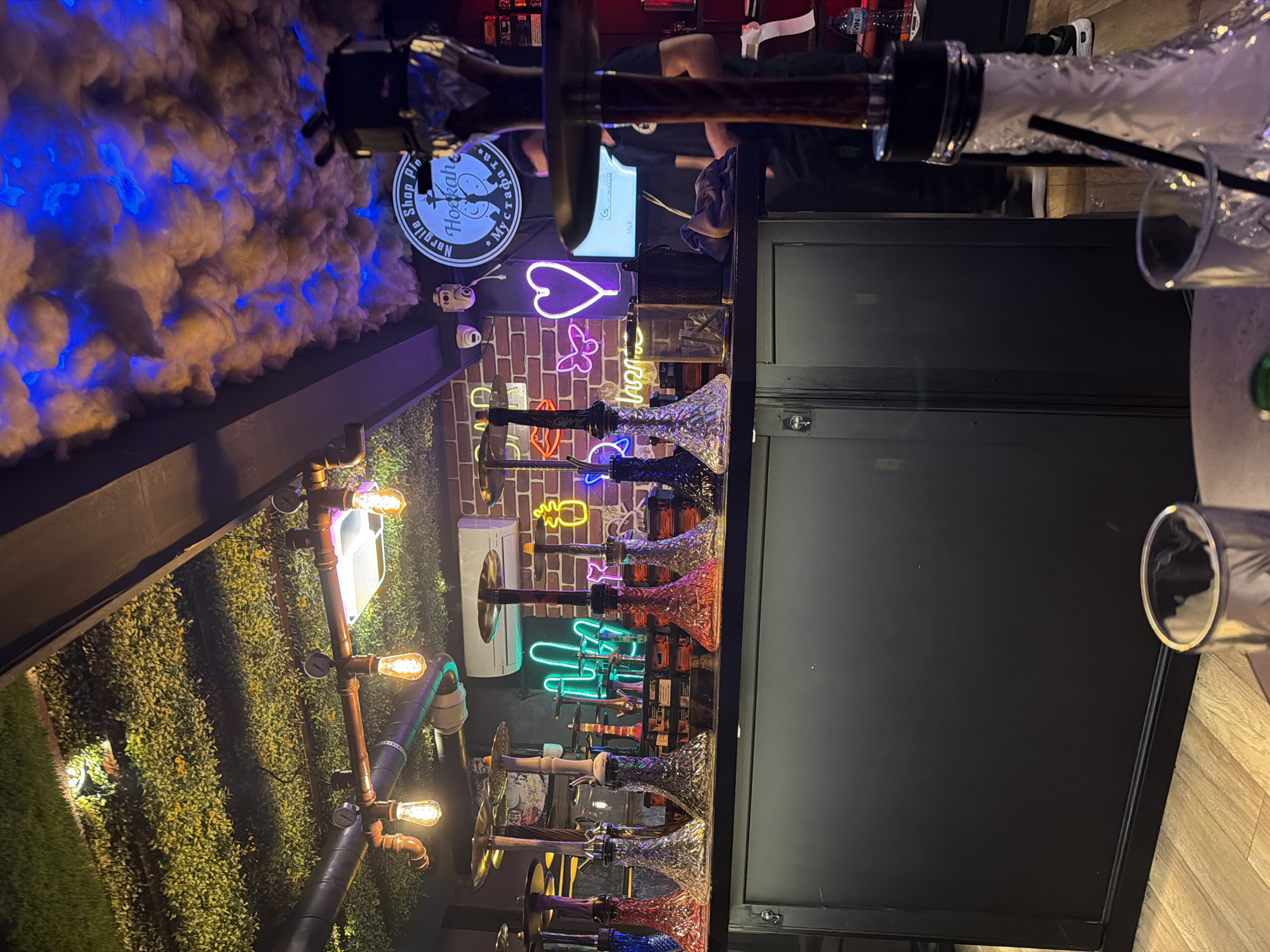 The image shows the interior of "Nargile Shop Plovdiv Myсtерии," a hookah bar located in Plovdiv, Bulgaria. The scene includes hookahs displayed against a brick wall decorated with neon signs, including a heart, butterfly, pineapple, and cactus. A structure with industrial-style lighting and artificial greenery is present. There's also a cloud-like decorative element with blue lighting. In the foreground, a hookah and a beverage glass are visible on a table with a wooden surface. People are partially visible in the background near a computer monitor. A black cabinet is adjacent to the display.