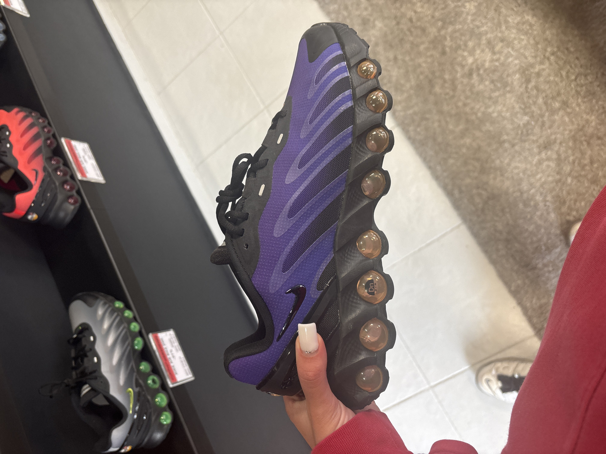 The image shows a person holding a black and purple Nike Air Max Plus sneaker with visible "Tn AIR" branding on the sole's air pods. The background features a display shelf with other sneakers, including a black and red pair and a black and grey pair. The floor is tiled, and a small portion of another person's shoe (white with black accents) is visible. The person holding the sneaker is wearing a red sweatshirt. The photo appears to be taken indoors, possibly in a shoe store, but there's no information to determine if it is in Gabrovo, Bulgaria.