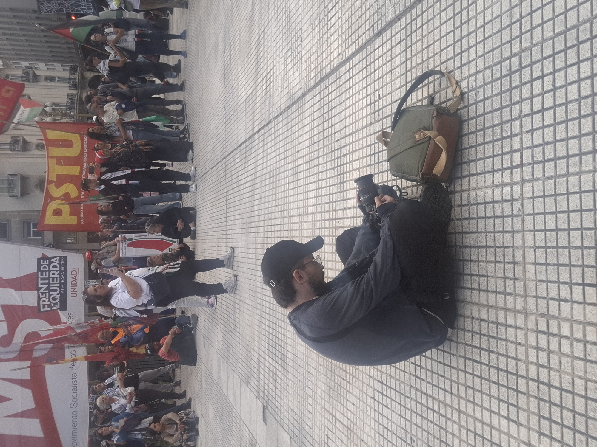 Here's a description of the scene:
The image shows a political protest taking place in Buenos Aires, Argentina. A crowd of people is gathered, holding banners and flags with socialist and leftist slogans. One large banner reads "FRENTE DE IZQUIERDA," and another features the acronym "PSTU".
A photographer is sitting on the tiled ground, taking pictures of the demonstration with a professional camera. A shoulder bag sits next to him. He is wearing a dark baseball cap and a long-sleeved shirt.