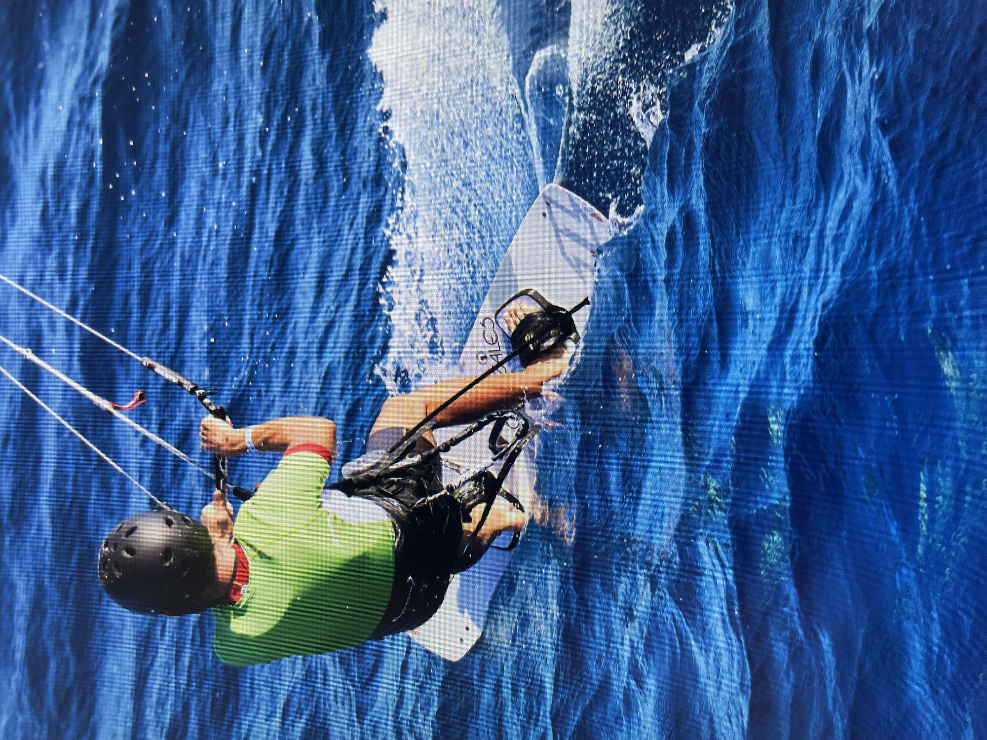 A man kiteboards on deep blue water, spraying a large wave to his right. He wears a black helmet, a lime-green and red short-sleeved shirt, dark shorts, and a black harness. He's gripping the kite's control bar with both hands, lines extending upwards from the bar. He is riding a white kiteboard with black bindings. The water is deep blue with subtle ripples and white spray. The location is Bacong, Philippines.