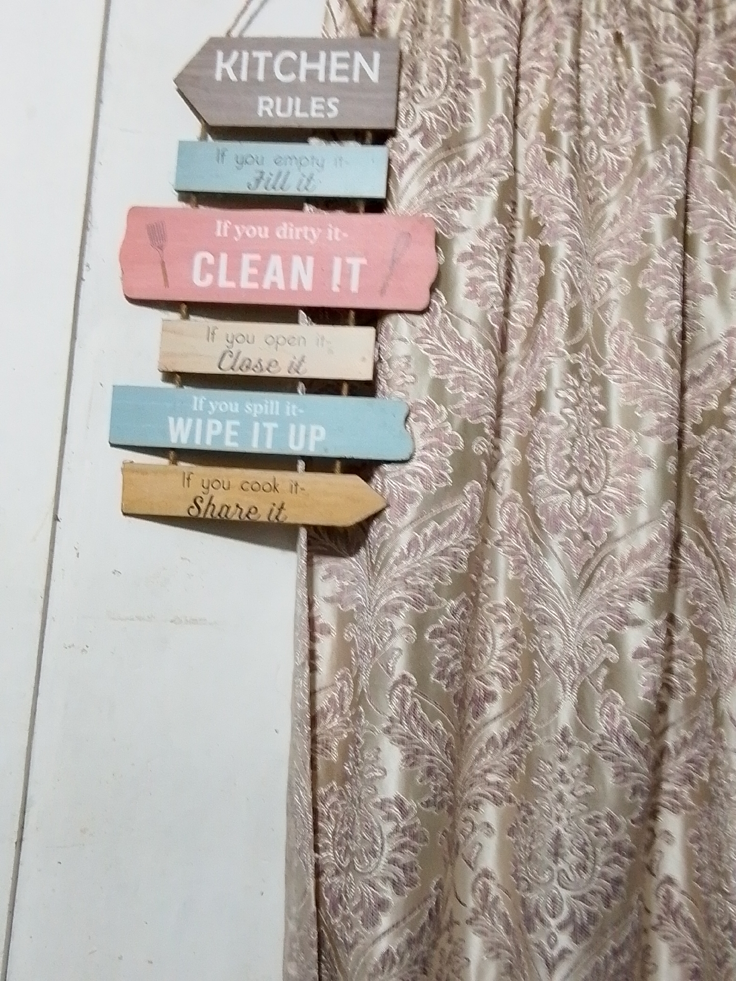 The media file displays an indoor scene, likely a kitchen or dining area, featuring a decorative "KITCHEN RULES" sign hanging on a light-colored wall. To the right, a rich, patterned curtain partially covers the view.
The sign is composed of six individual, horizontally stacked wooden planks, each shaped like an arrow pointing alternately left and right. The top plank, colored gray, reads "KITCHEN RULES". Below it, the rules are listed:
- On a light blue plank: "If you empty it- Fill it"
- On a pink plank, accompanied by a whisk icon: "If you dirty it- CLEAN IT"
- On a natural wood plank: "If you open it- Close it"
- On another light blue plank: "If you spill it- WIPE IT UP"
- On a final natural wood plank: "If you cook it- Share it"
The wall appears to be white or off-white, showing minimal texture or marks. The curtain, partially visible on the right, is made of a heavy fabric with an intricate, possibly damask or brocade, pattern. The background color of the curtain is a light cream or beige, with the ornate patterns rendered in a contrasting dusty rose or mauve hue. The curtain is gathered, indicating it's drawn across a window or opening.
The lighting suggests an indoor setting during the daytime. No people or specific activities are depicted, focusing instead on the home decor element. The overall impression is one of a domestic space, emphasizing order and shared responsibilities within the kitchen. While the context mentions Davao City, Philippines, no specific visual cues in the image itself denote this location.