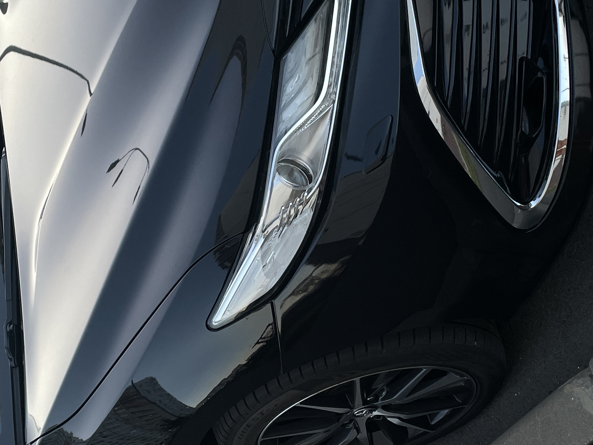 The image is a close-up, low-angle exterior shot of the front quarter panel, headlight, wheel, and grille of a black car. The car is parked on a paved surface, possibly a street or parking lot. The environment appears to be outdoors, with hints of a building or fence visible in the reflection on the car's surface. The lighting suggests it may be daytime or early evening, with reflections of the sky and possibly streetlights or other urban elements on the car's polished black paint. The image does not contain any information that allows for its location to be identified as Rostov-on-Don, Russia.