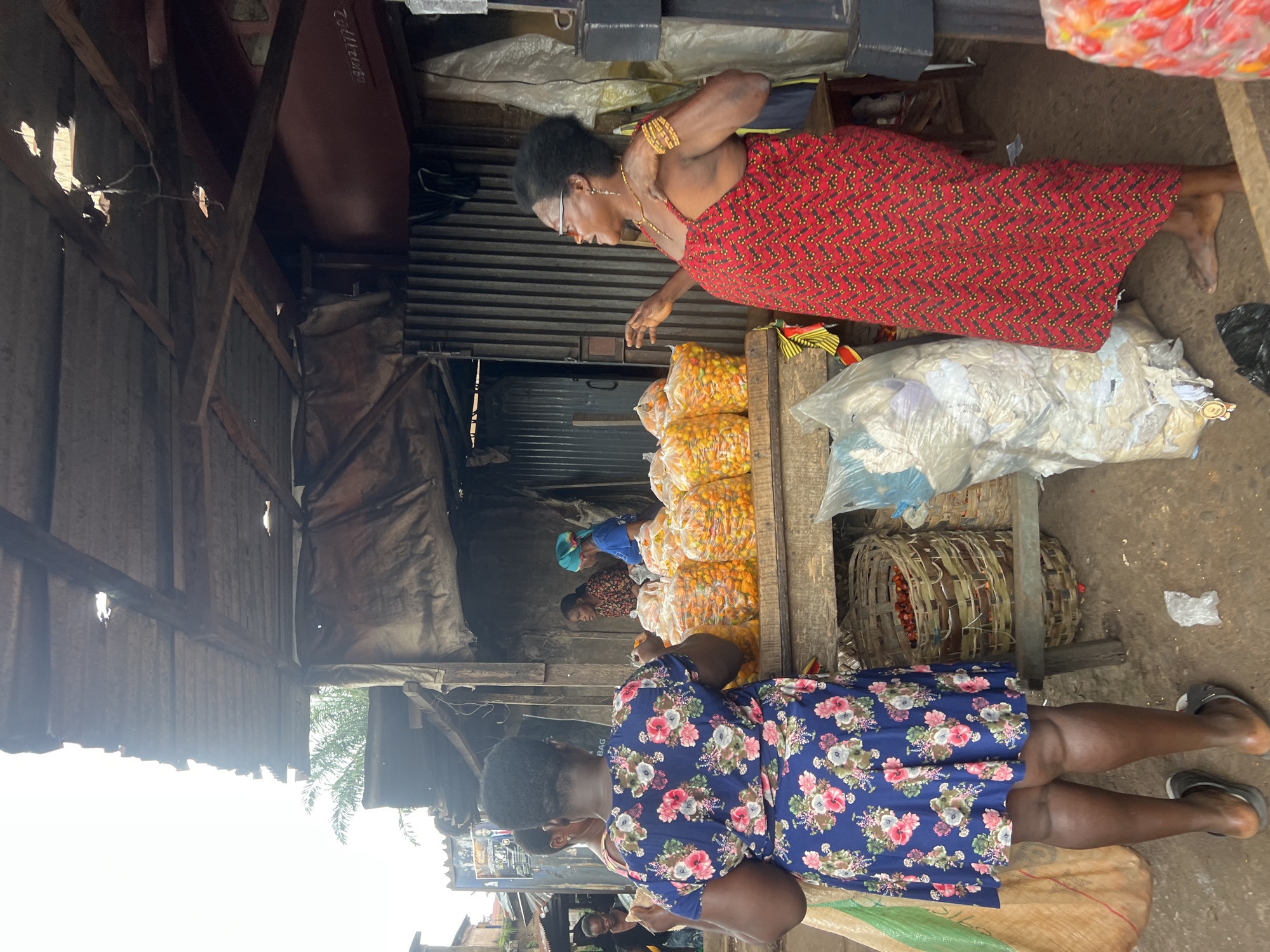 The image depicts a bustling outdoor market scene in Nkpor, Nigeria. In the foreground, two women are engaged in a transaction. The woman on the left, seen from behind, wears a blue floral dress and stands with her back to the camera. To her right, a woman in a red, patterned dress with spaghetti straps gestures with her hands, appearing to be speaking animatedly. In front of them, a wooden stall is laden with numerous clear plastic bags filled with small, yellow-orange fruits, likely peppers or citrus. Baskets containing red produce are also visible below the stall. The market structure itself is rustic, with a corrugated metal roof supported by wooden beams, and tarpaulin sheeting providing shade. In the background, other marketgoers and stalls are discernible, suggesting a vibrant commercial environment. The ground is dirt and scattered with some debris.