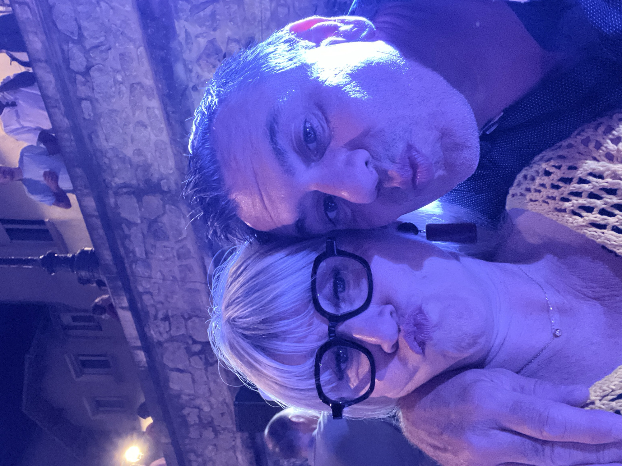 A selfie is taken by a couple in the city of Saint-Galmier, France, at night. The couple is posing for the photo, with both of them puckering their lips. They are dressed in casual clothing. The woman on the left is wearing glasses and a yellow knitted top, and a necklace. The man on the right is wearing a dark shirt. In the background, there are stone walls and buildings, as well as other people and lights. The scene appears to be outdoors, possibly at a festival or event, indicated by the ambient lighting and the presence of multiple people.
