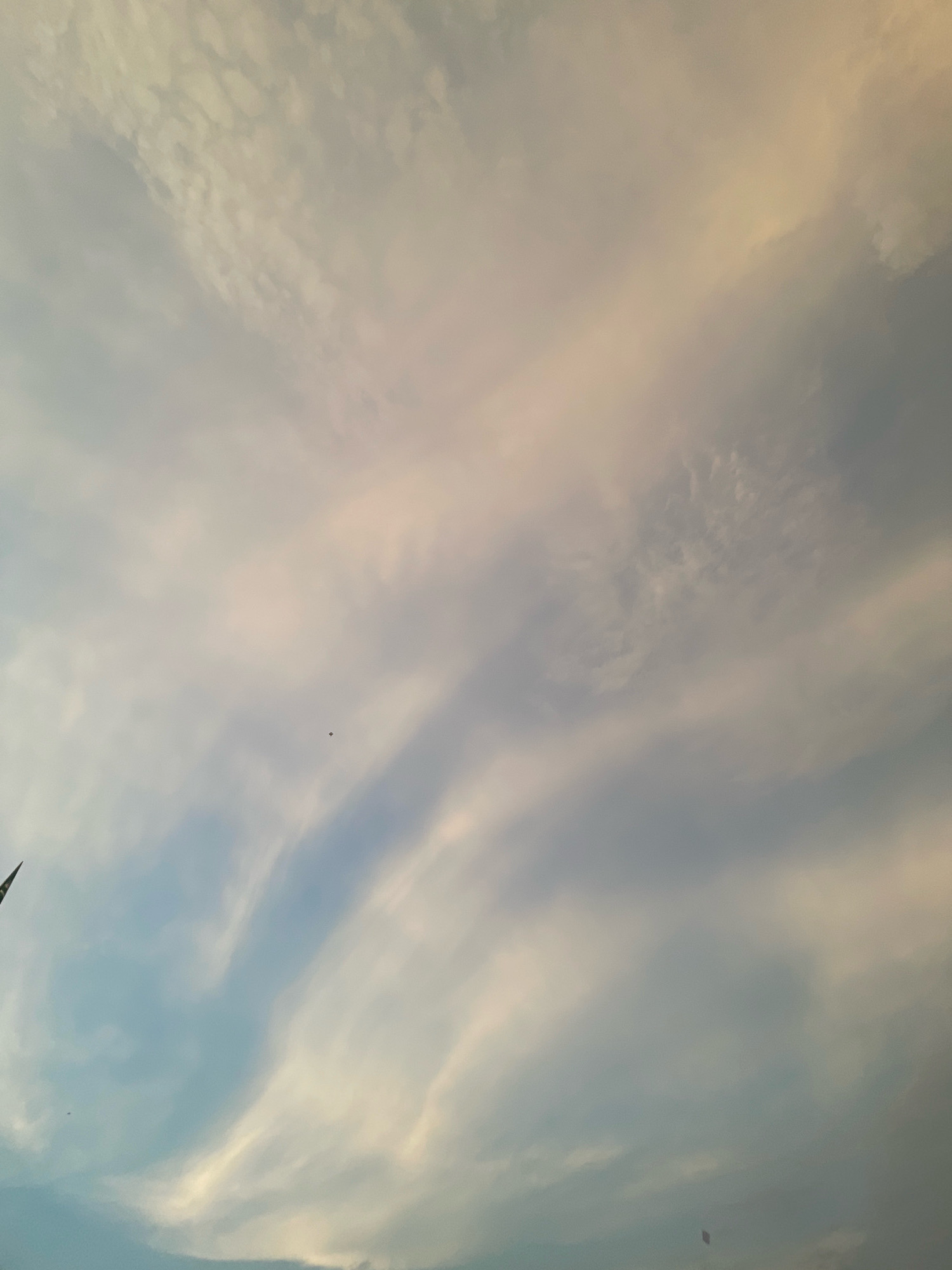 The image is a sky view, likely captured in Bojhi, India. The sky is filled with a mix of light blue and pale yellow clouds, suggesting it might be either early morning or late afternoon. The clouds are wispy and streaky, with some areas appearing denser and more textured than others. There are at least two kites visible in the sky. One kite, small and dark, is positioned in the middle left of the frame. Another kite, also dark, is visible in the bottom right corner. A portion of what appears to be a flag or tall structure is present on the far left edge of the image, partially obscuring the sky. The overall impression is of a serene, open sky with signs of recreational activity.
