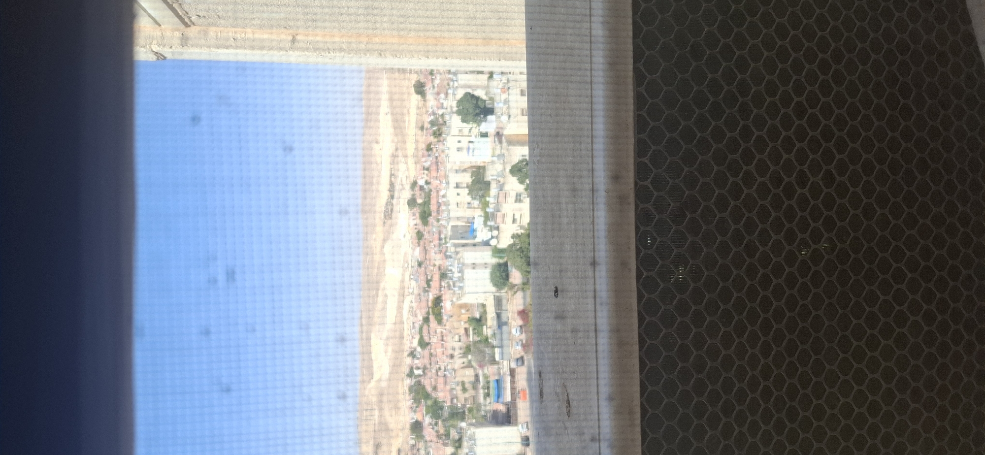 The image is a photograph taken from an indoor vantage point looking out a window. Through a screened window, a cityscape is visible. The cityscape consists of multiple buildings, varying in height and architectural style, with many featuring reddish-brown tile roofs, common in Israel. Trees are interspersed throughout the urban area. In the background, a barren, hilly landscape under a bright blue sky suggests a desert environment, consistent with the location of Dimona, Israel. A rough, textured surface, possibly a concrete wall, is visible on the right side of the frame. The bottom portion of the image shows a dark, hexagonal patterned mesh, likely part of the window screen or an air conditioning unit. The overall scene depicts a residential area in Dimona, Israel, under clear daytime conditions.