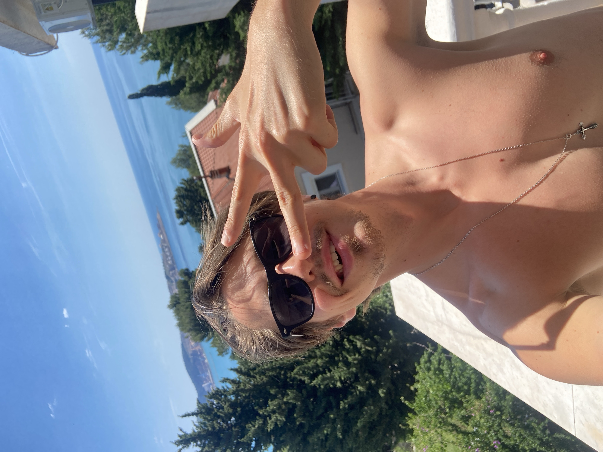 A shirtless Caucasian male is shown from a low angle, with his body nearly parallel to the top of the frame. He is wearing dark sunglasses and a silver necklace with a cross pendant. His right hand is raised, with his index and middle fingers extended, forming a 'V' shape over his left eye. Behind him, the Adriatic Sea stretches into the distance under a bright blue sky. Green trees and a building with a red tile roof are also visible. Based on the landscape, the location is likely Šušanj, Montenegro.