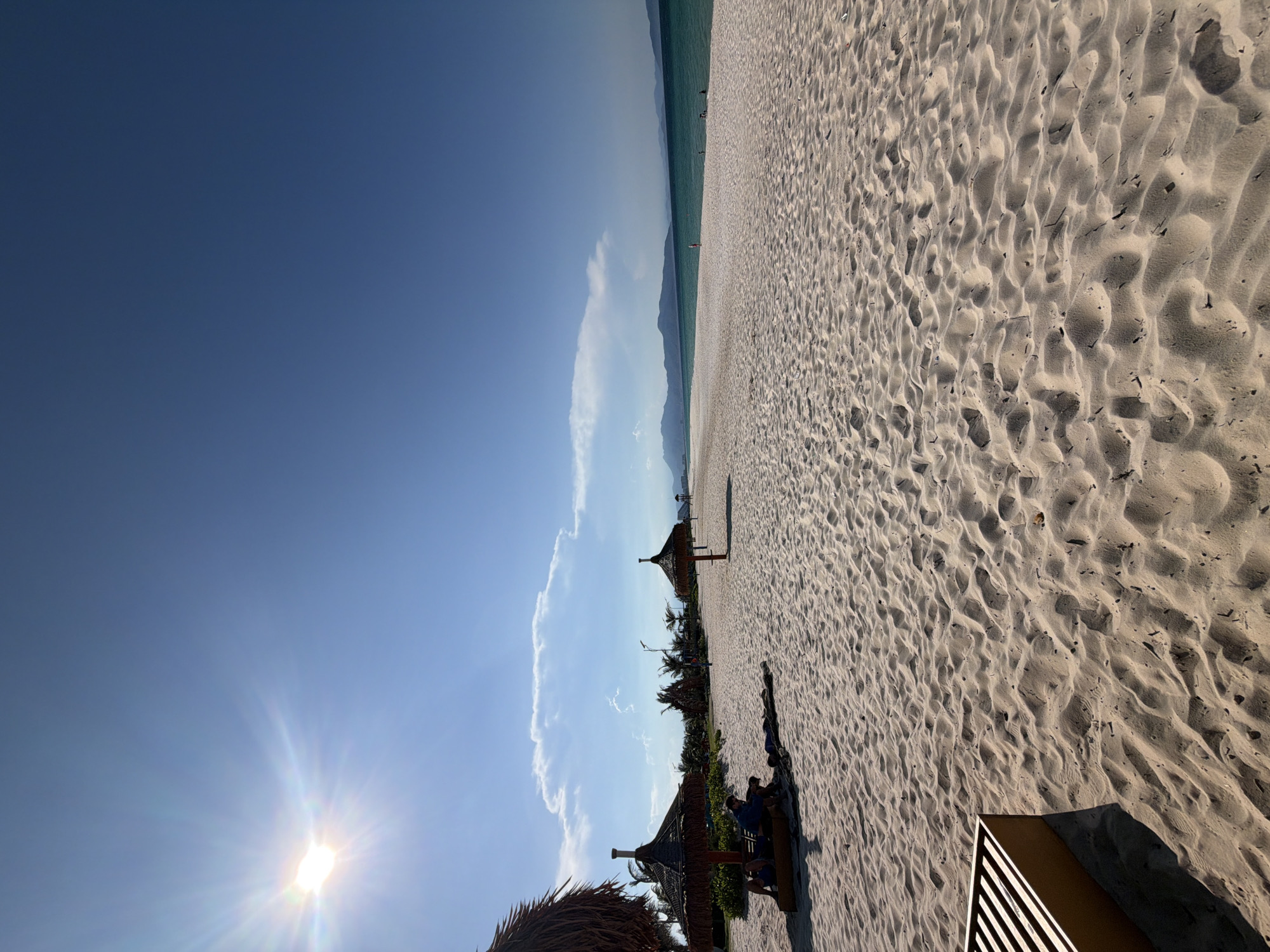 The image depicts a wide, sandy beach under a clear blue sky with the sun shining brightly in the upper left corner. Several thatched-roof umbrellas are scattered across the sand, providing shade. In the foreground, to the left, two individuals are seated on a bench under one of these umbrellas, with another person lying down nearby. Further down the beach, more umbrellas are visible, and the turquoise ocean stretches to the horizon, where it meets a distant, hazy mountain range. The sand is textured with ripples, indicating wind or water movement. This scene is characteristic of a tropical beach location, likely in or near Bac Cam Ranh Ward, Vietnam, known for its coastal resorts.