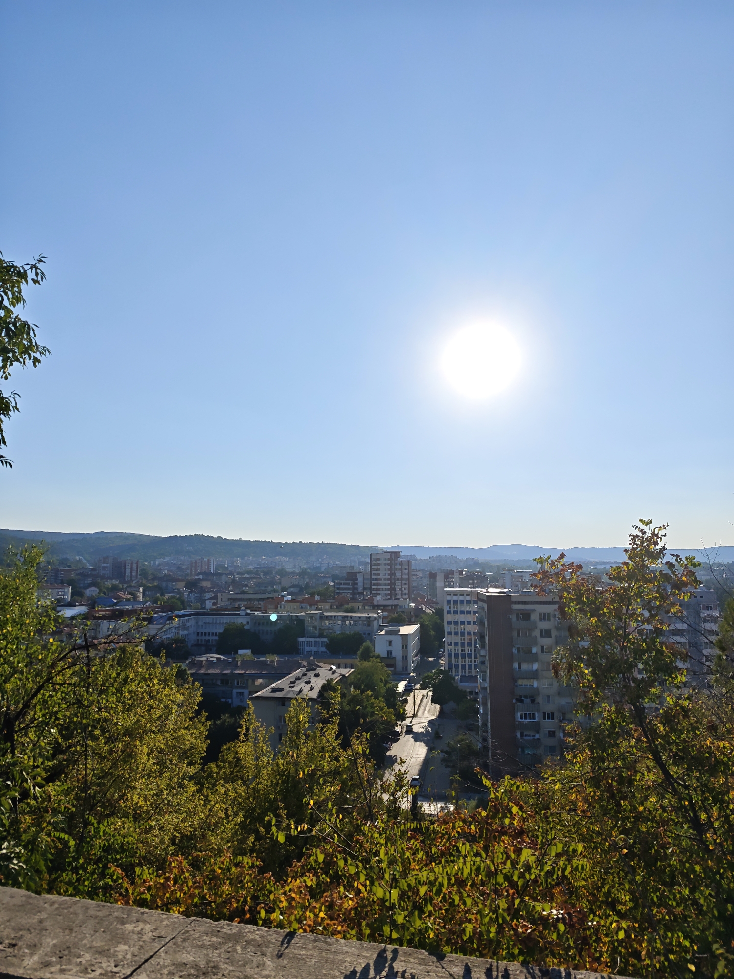 This is a vertical, medium-distance, high-angle, daytime, outdoor, photograph taken from a vantage point overlooking the city of Lovech, Bulgaria. The sun is high in the clear blue sky, positioned slightly right of the center, and appears as a bright, luminous disk. In the foreground, there is a concrete structure, possibly a wall or parapet, with some shadows cast upon it. Lush green and autumnal foliage, with some yellow and orange leaves, frames the lower portion of the image.
Beyond the foliage, the cityscape of Lovech unfolds. A road with several cars is visible, winding through the urban area. Various buildings, including multi-story residential blocks, some appearing modern with balconies, and others older with tiled roofs, are scattered across the landscape. The buildings are of different heights and architectural styles. In the distance, rolling hills form the horizon, adding depth to the scene. The overall atmosphere is one of a bright, sunny day in an urban setting with natural elements.
