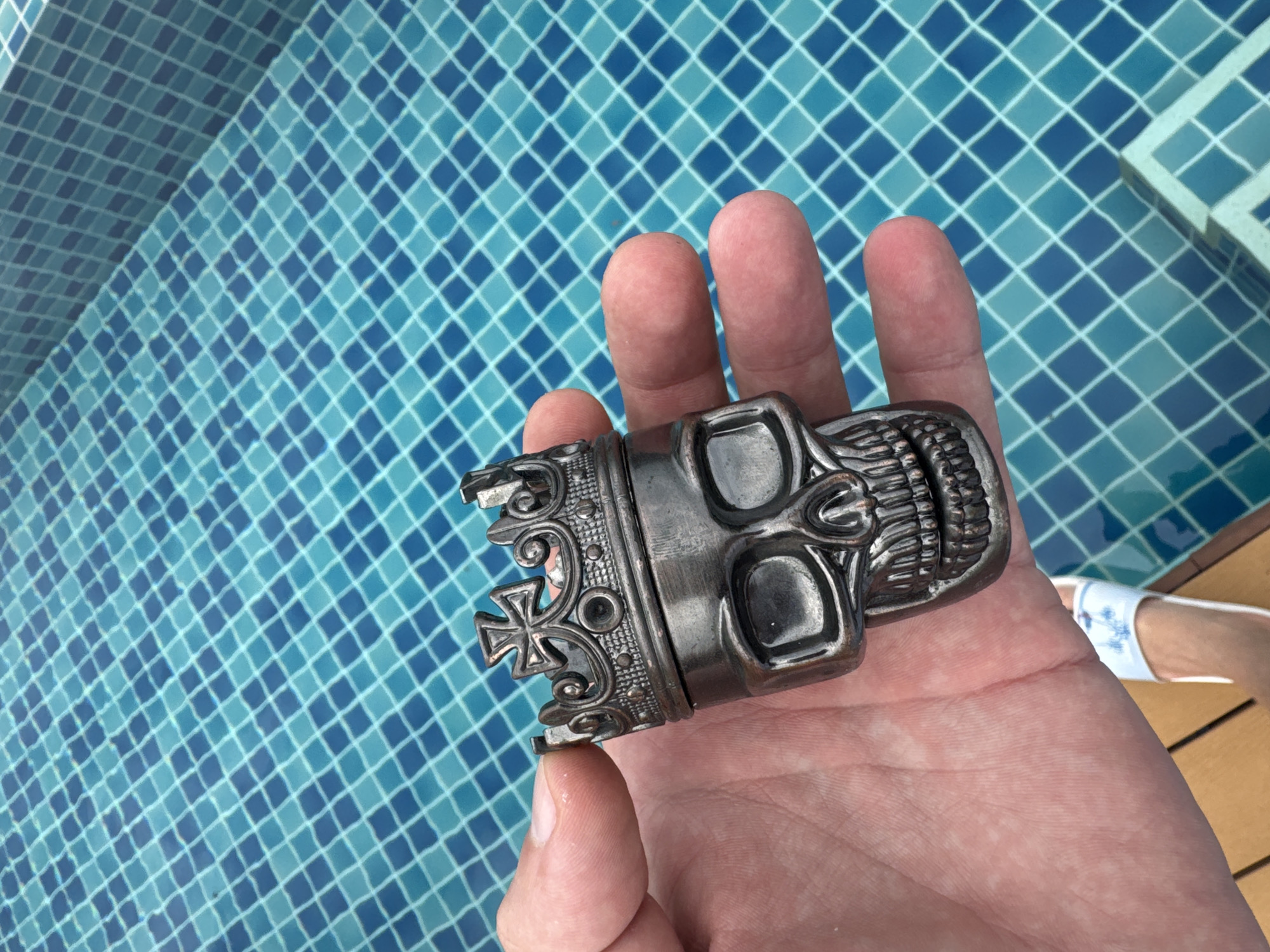 An overhead shot shows a person's hand holding a metal skull with a crown, positioned above a blue tiled swimming pool. The skull is detailed with a crown, eye sockets, and teeth. The pool is partially visible in the background and foreground, showing the mosaic of blue tiles. The scene appears to be at a resort or private residence in Rawai, Thailand.
