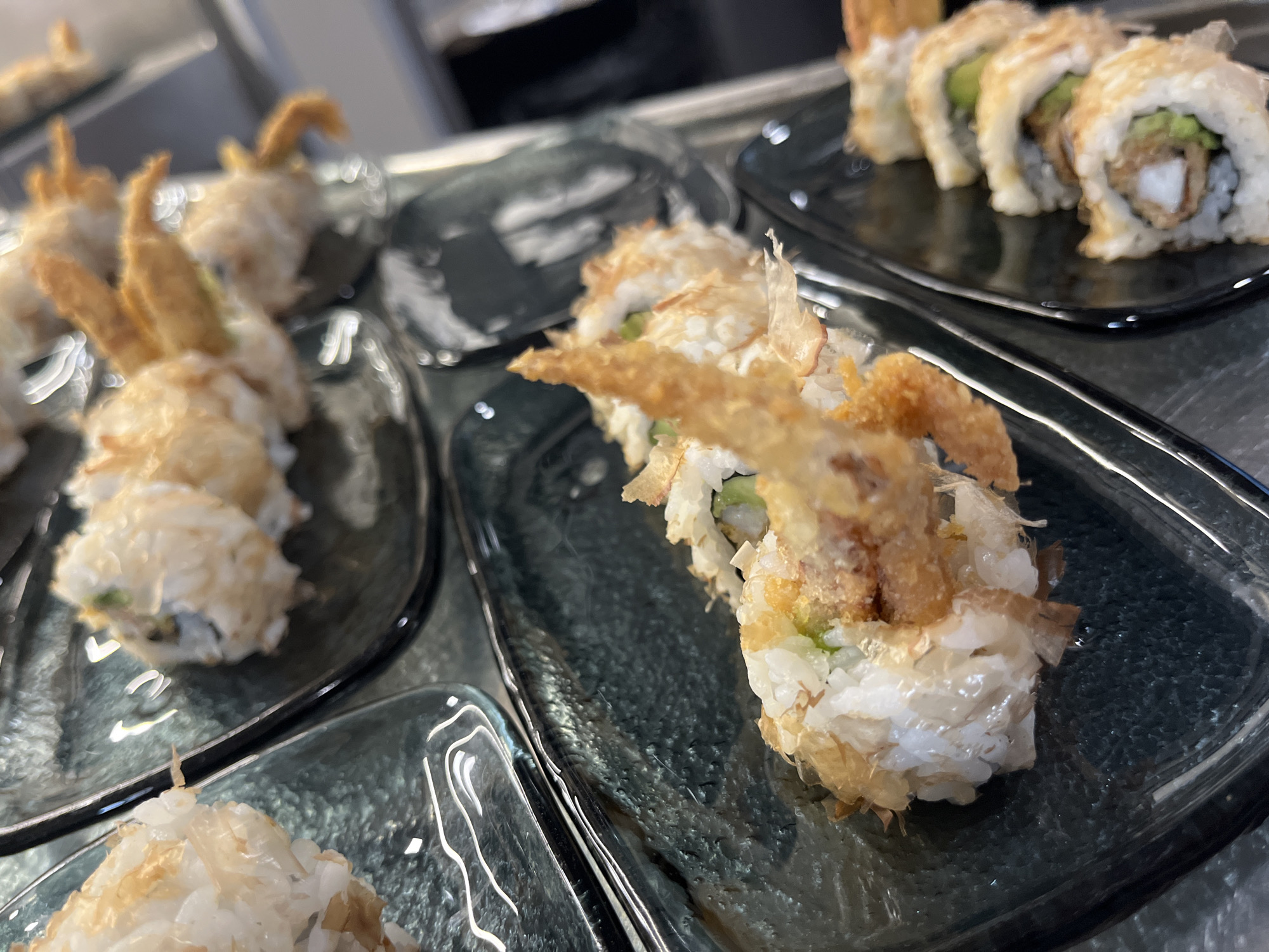 Several plates of sushi are arranged on a glass surface. The sushi rolls are of two distinct types: one variety is topped with what appears to be fried crab legs and coated with shaved dried bonito flakes, and the other type appears to contain fried shrimp and avocado. The plates holding the sushi are dark-colored with a square shape, and the glass surface beneath them suggests a display case or countertop. The image does not provide information regarding people, actions, interactions, or the location in Sofia, Bulgaria.