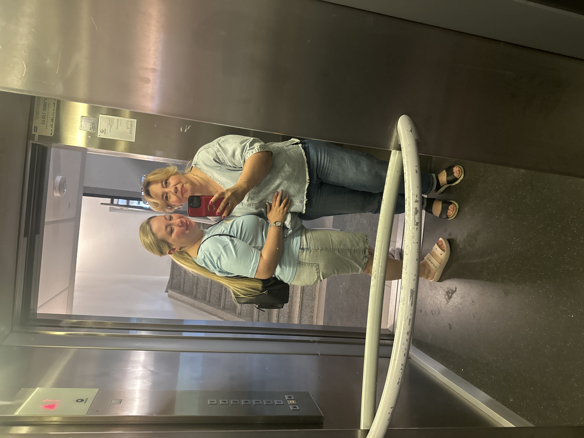 Two women are reflected in a mirrored elevator. The woman in the foreground, with blonde hair in a ponytail, is wearing a light blue t-shirt and a denim skirt. She is holding a red cell phone and taking a selfie. The woman behind her, who is also reflected, has blonde hair and is wearing a light blue denim shirt and blue jeans. Both women are smiling.
The elevator has stainless steel walls and a gray floor with small flecks. A white handrail runs along the right side of the elevator. In the background, visible through the elevator doors, is a staircase. The time of day and weather are not discernible from the image.
On the elevator wall, there are several labels. One label is white with black text and appears to be an information or safety notice. Another label is white with black text and a logo, possibly a service provider. An illuminated panel with buttons is visible on the elevator wall, showing a red "5" and an arrow, indicating the elevator is on the fifth floor.
Visible text on the labels includes "0800 120 03" and "D Helburg B12 B3".