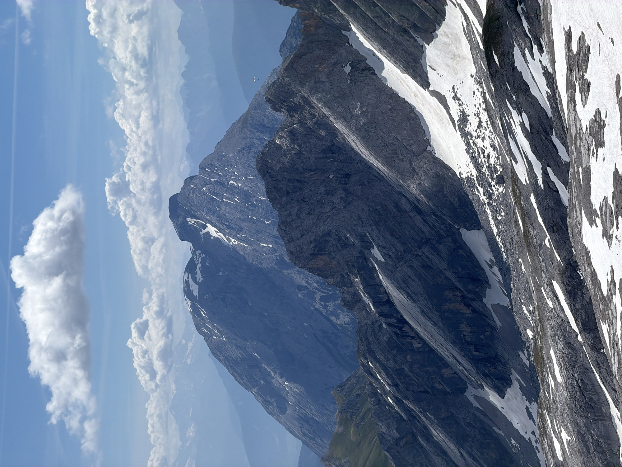 The image depicts a majestic mountain range under a partly cloudy sky. The foreground is dominated by jagged, rocky peaks with patches of snow scattered across their slopes. These slopes are steep and uneven, with visible scree and rock formations. Further into the background, larger, more imposing mountains rise, some with snow-capped summits. The sky is a vibrant blue, with scattered white cumulus clouds casting shadows on the landscape. In the mid-ground, a lone paraglider can be seen, a small white speck against the vastness of the mountains. The overall impression is one of rugged, natural beauty and a sense of scale. The scene is consistent with a view from a high vantage point in the Bavarian Alps, such as the Zugspitze, Germany's highest peak, located near Garmisch-Partenkirchen.