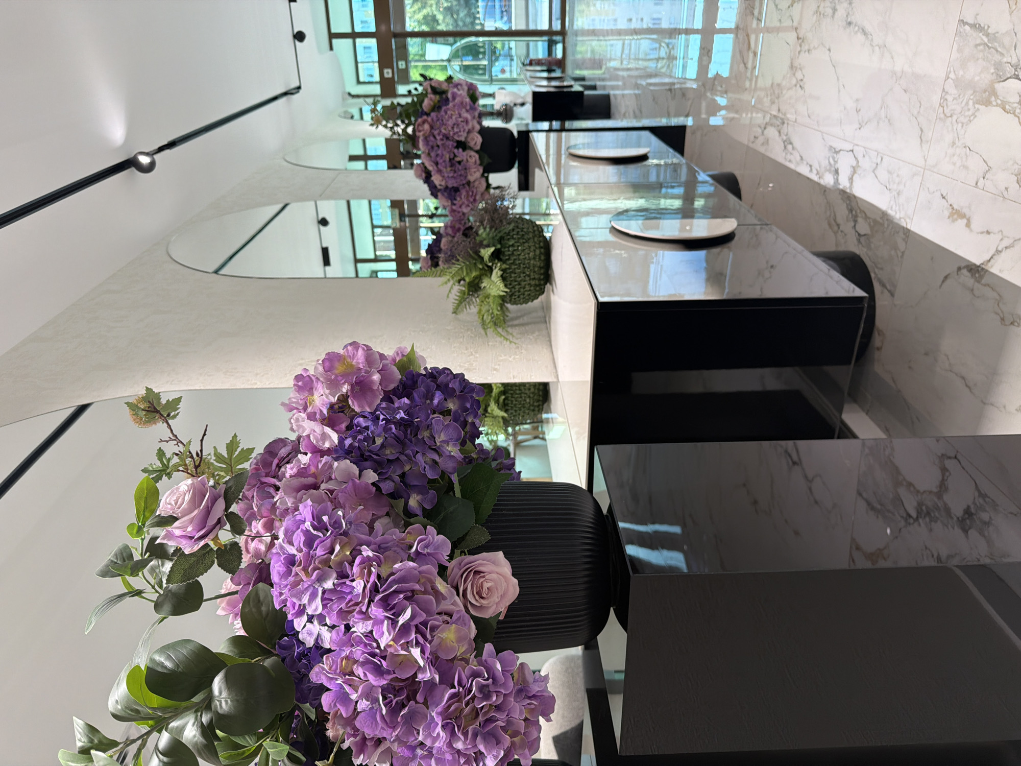 A modern restaurant interior features tables set for dining, arranged in a row along a wall. The tables have black, reflective surfaces and are set with white plates. Floral arrangements with purple hydrangeas and roses in black vases decorate the tables. A wall with marble-like tiles forms a backdrop. Mirrors adorn the wall above the tables. Light fixtures are visible on the ceiling. The windows reflect an outdoor scene with trees and buildings. The scene suggests a clean, contemporary design aesthetic, likely in a commercial establishment. There is no definitive evidence of Blagoevgrad, Bulgaria from this image.